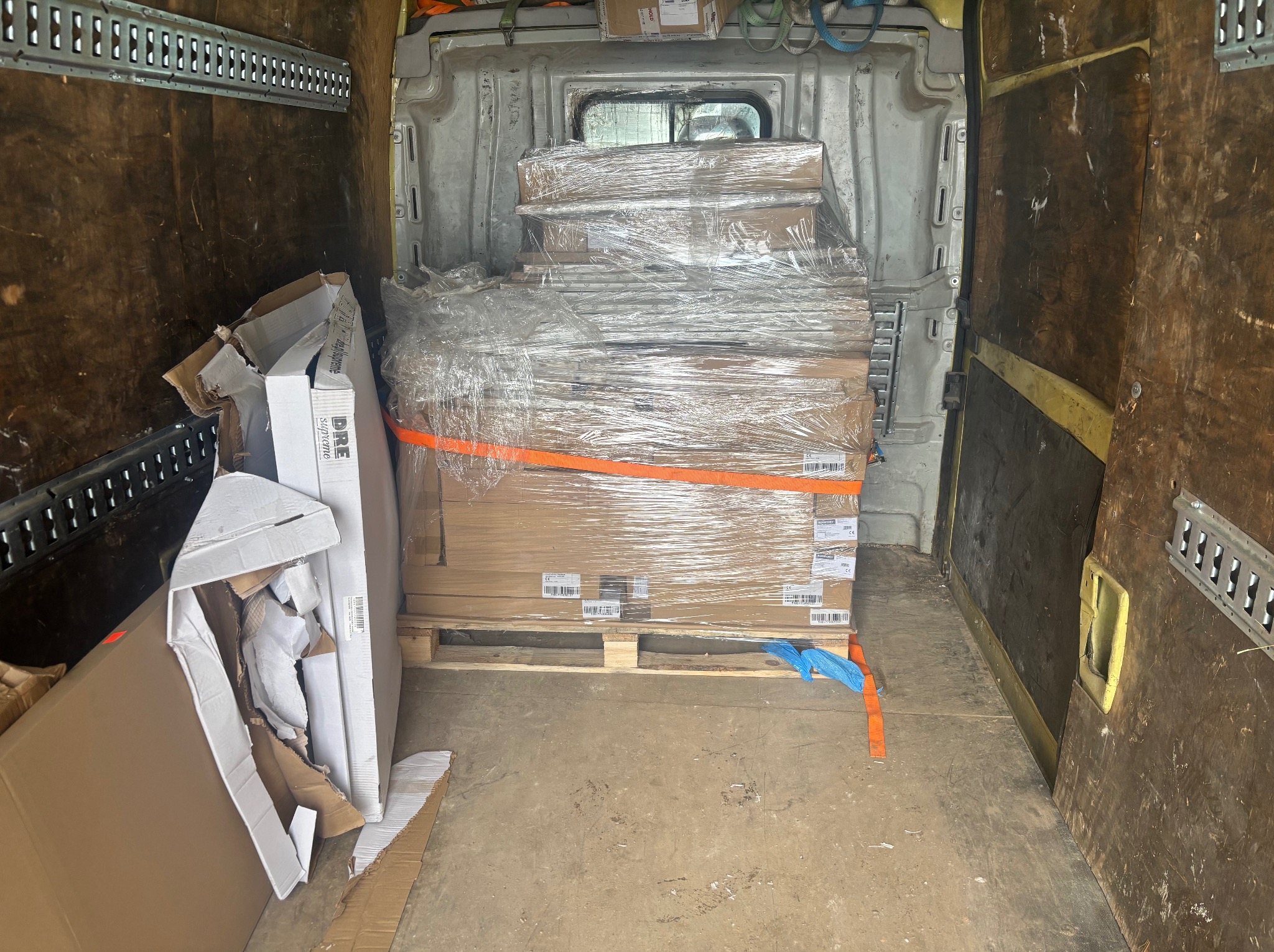 Kartonowe pudła na palecie, owinięte folią stretch i zabezpieczone pasem transportowym wewnątrz brudnej przestrzeni ładunkowej busa. Częściowo rozpakowane kartony obok.