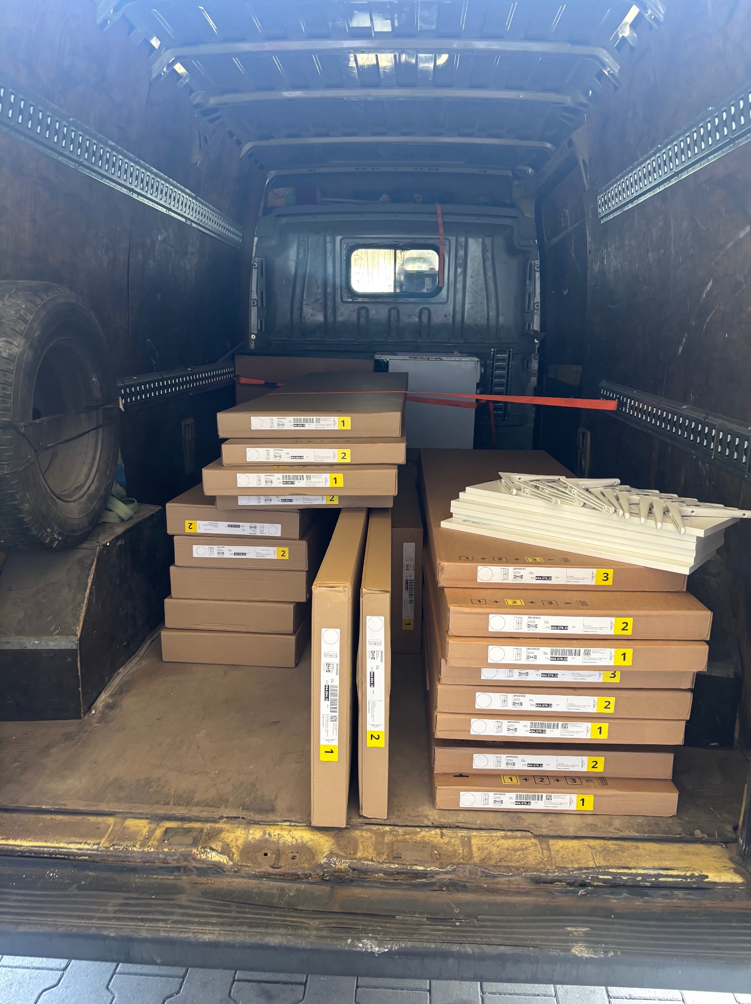 Wnętrze busa wypełnione kartonami z meblami IKEA, zabezpieczonymi pasami transportowymi. Widoczne koło zapasowe i metalowe ściany pojazdu. Perspektywa z tyłu pojazdu.