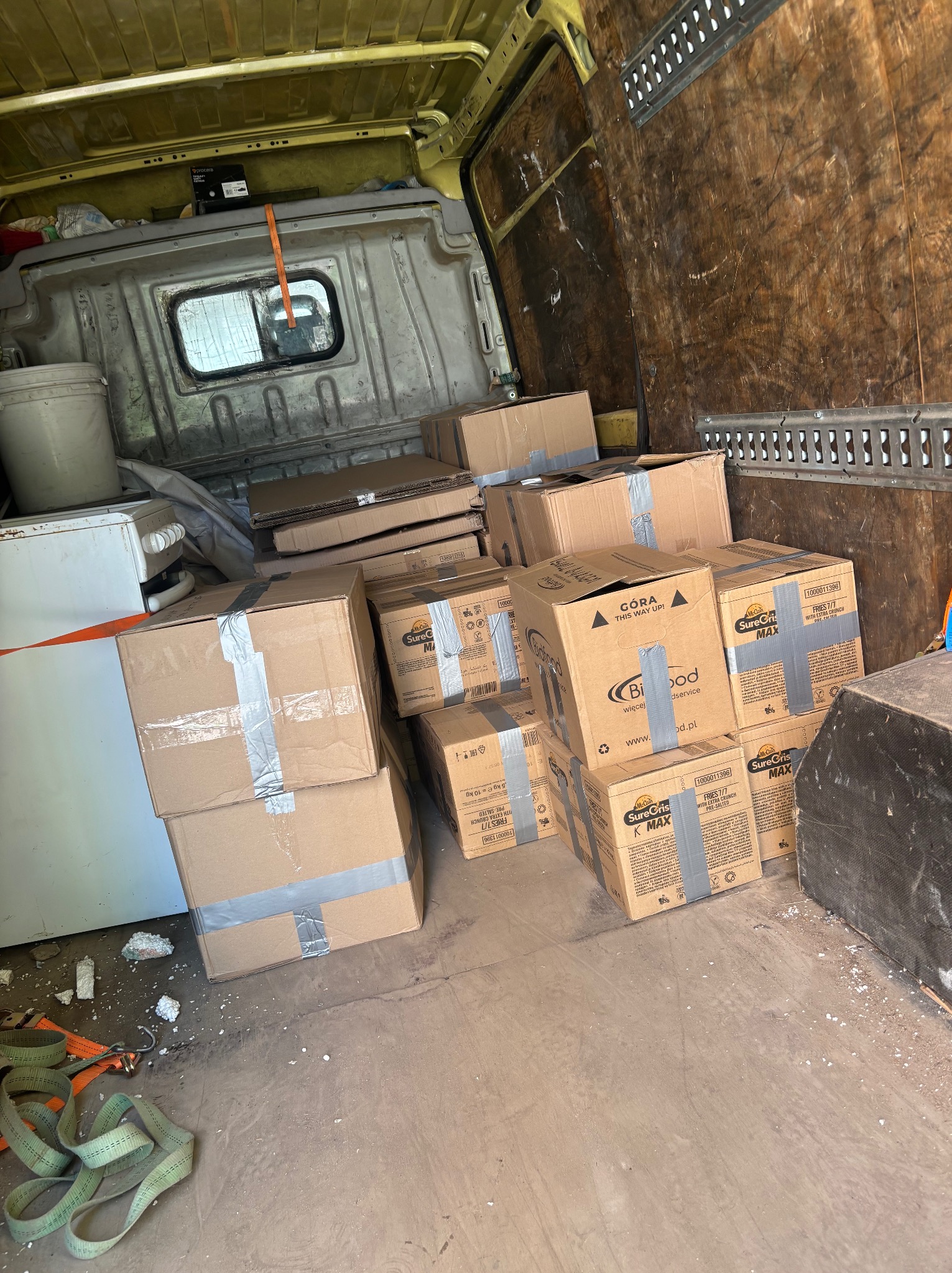 Wnętrze busa wypełnione kartonami, zabezpieczonymi taśmą, gotowymi do transportu. Widoczne pasy mocujące i elementy wyposażenia pojazdu.