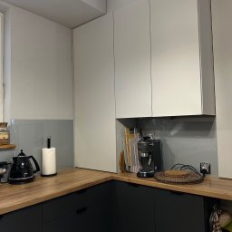 CZYSTY DOM - Nowoczesna kuchnia z drewnianym blatem, czarnymi szafkami i białymi szafkami wiszącymi. Na blacie czajnik, ekspres do kawy i akcesoria kuchenne. Szklany panel nad blatem.
