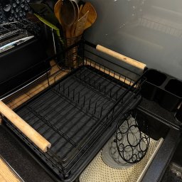 CZYSTY DOM - Czarny ociekacz na naczynia z drewnianymi uchwytami, umieszczony na czarnym blacie kuchennym obok zlewu. W tle przybory kuchenne i szara ściana z dekoracyjnym wzorem.