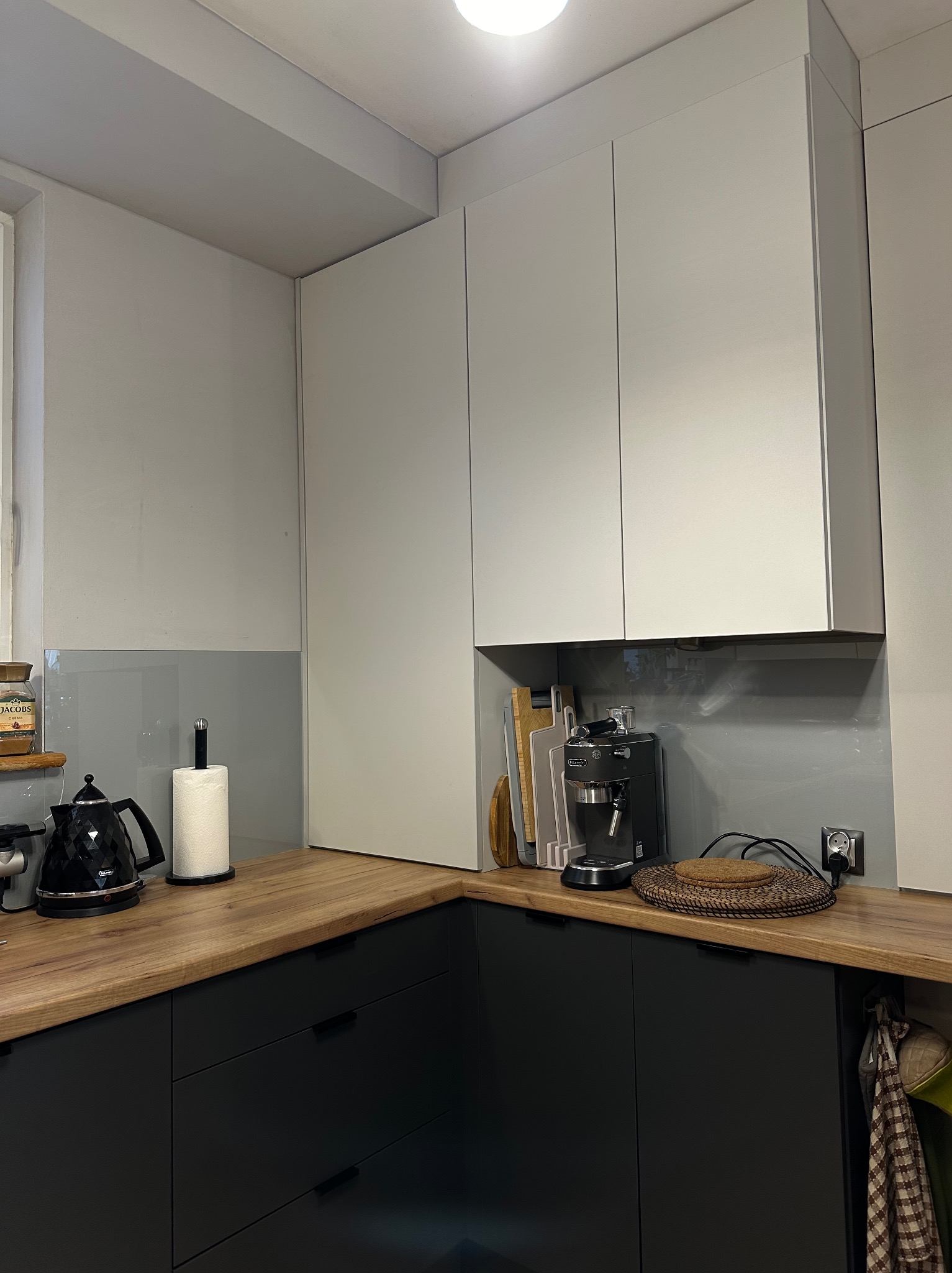 Nowoczesna kuchnia z drewnianym blatem, czarnymi szafkami i białymi szafkami wiszącymi. Na blacie czajnik, ekspres do kawy i akcesoria kuchenne. Szklany panel nad blatem.