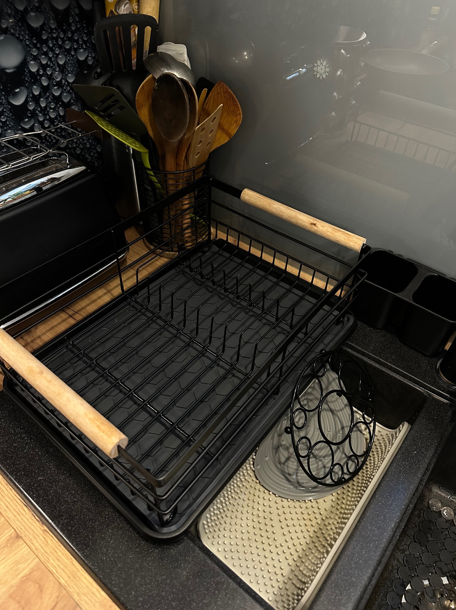 Czarny ociekacz na naczynia z drewnianymi uchwytami, umieszczony na czarnym blacie kuchennym obok zlewu. W tle przybory kuchenne i szara ściana z dekoracyjnym wzorem.