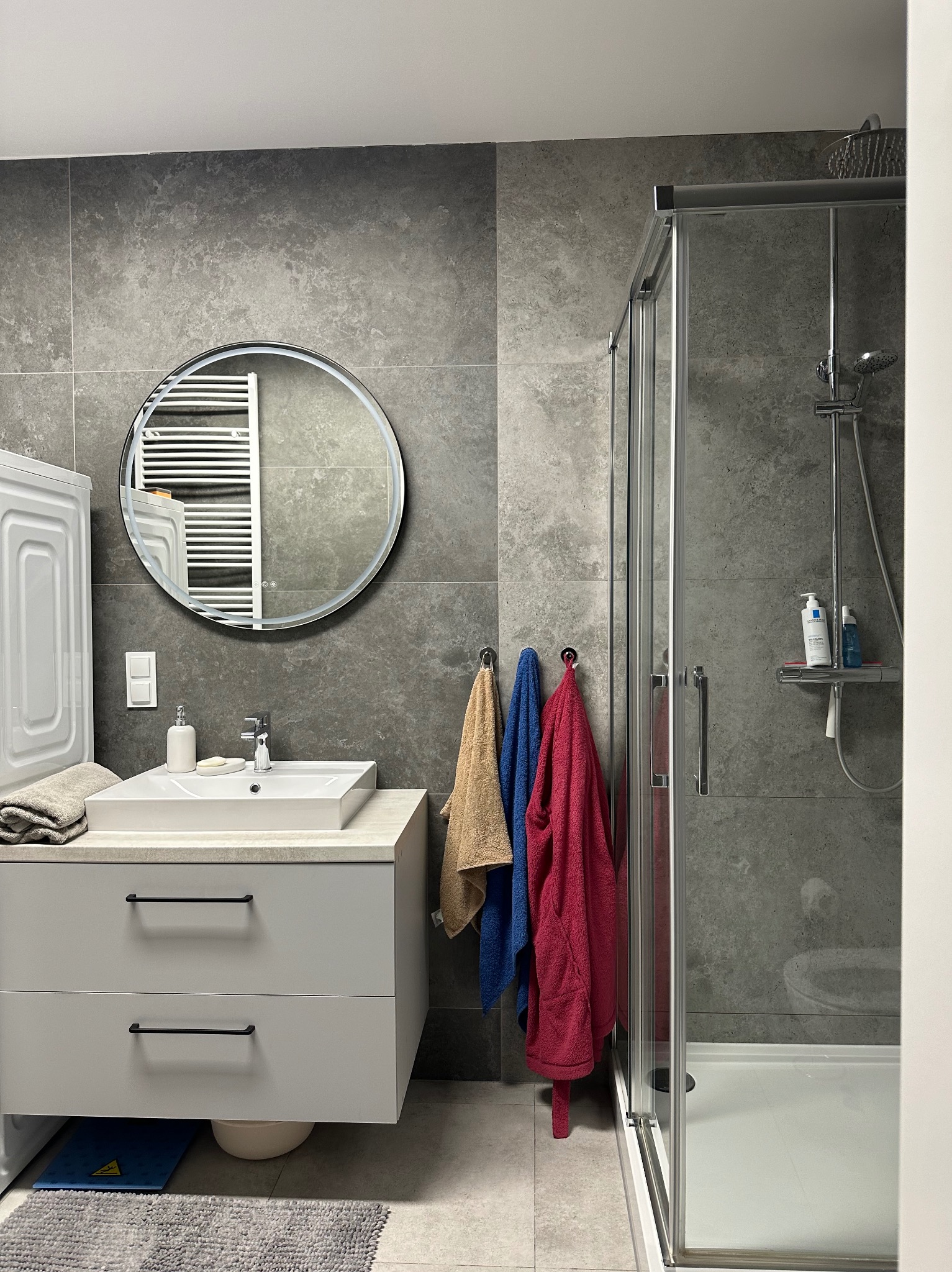 Nowoczesna łazienka z szarą armaturą, okrągłym lustrem z podświetleniem LED, kabiną prysznicową i wiszącą szafką pod umywalką. Widoczne ręczniki i pralka.
