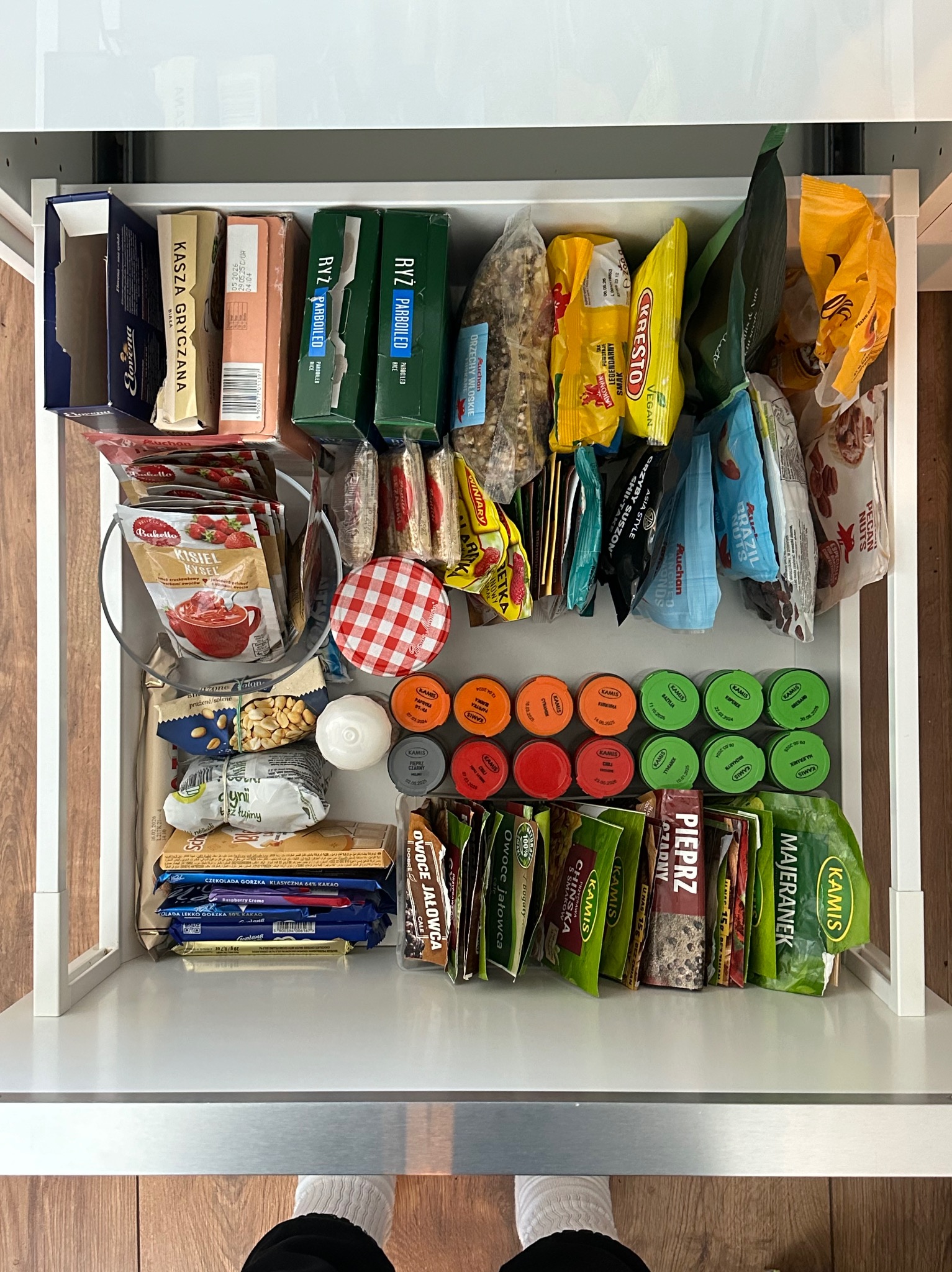 Pełna szuflada z zapasami: ryż, kasza, orzechy, przyprawy Kamisa, słodycze i kisiel w proszku, wszystko starannie ułożone w białej szufladzie.