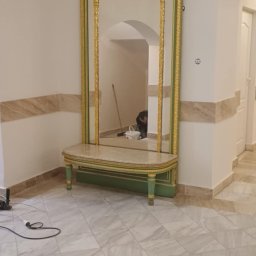 Usługi budowlane - Hall z dużym, ozdobnym lustrem w złotej ramie, stojącym na marmurowej konsoli. W lustrze odbija się osoba pracująca przy ścianie. Podłoga wyłożona marmurowymi płytkami.