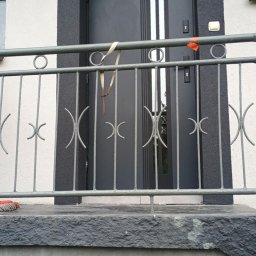 Usługi budowlane - Elewacja domu z ciemną ramą wokół drzwi wejściowych i ozdobną, metalową barierką. Widoczne wykończenie elewacji i detale architektoniczne.