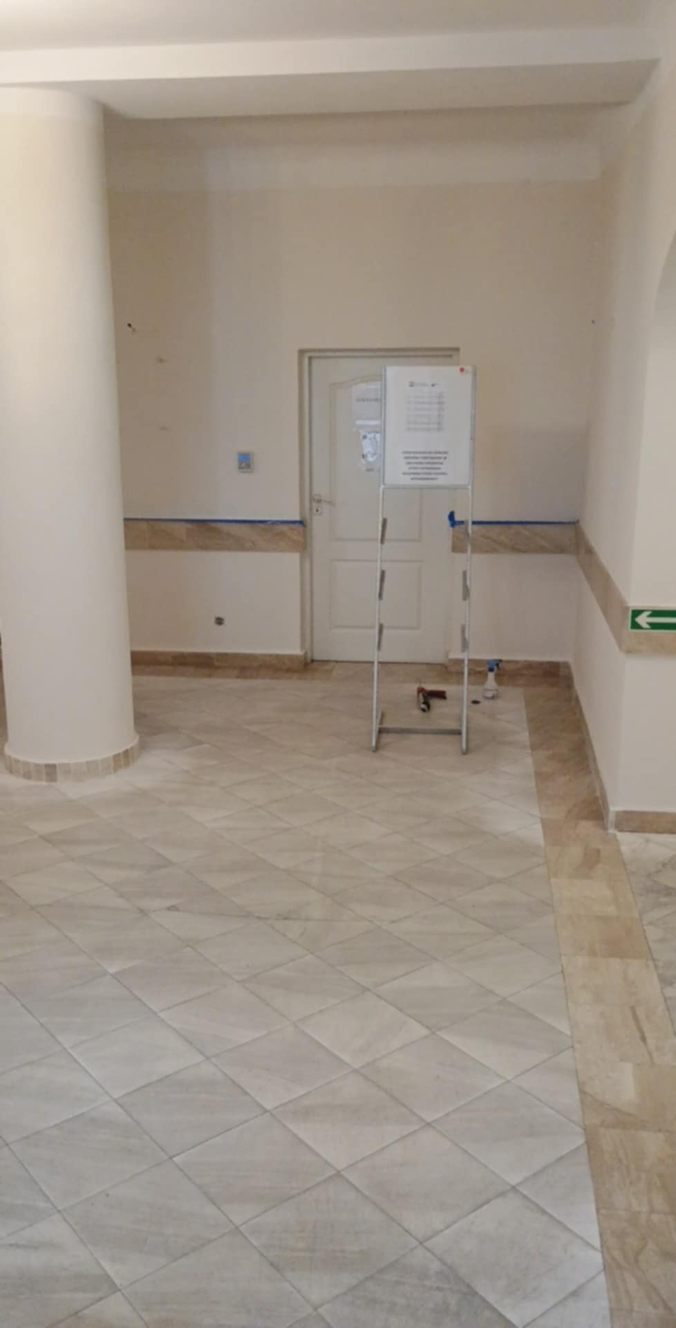 Jasne wnętrze z marmurową podłogą w romby i kolumną. Widoczna tablica informacyjna, listwy przypodłogowe i drzwi. Neutralna kolorystyka.