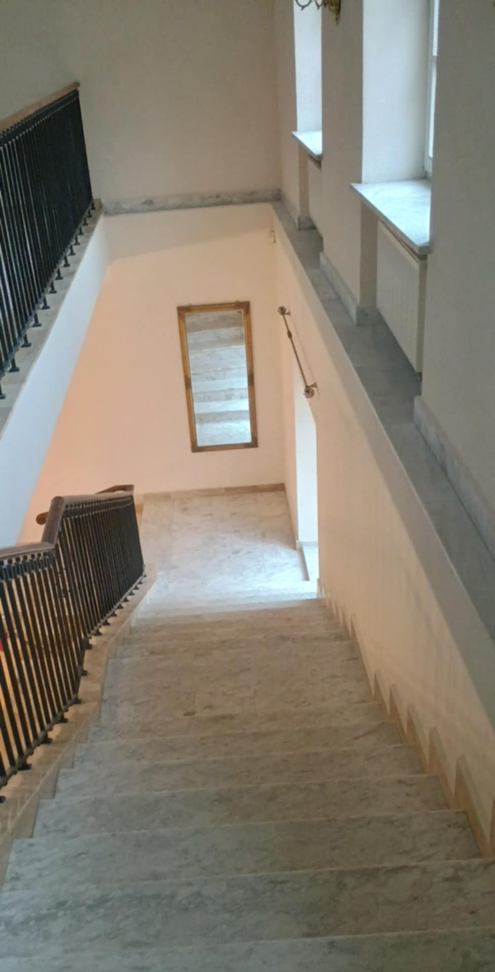 Marmurowe schody z kutą balustradą w kamienicy w Siedlcach. Na ścianie lustro w złotej ramie. Widok z góry na dół, jasne wnętrze, naturalne światło.