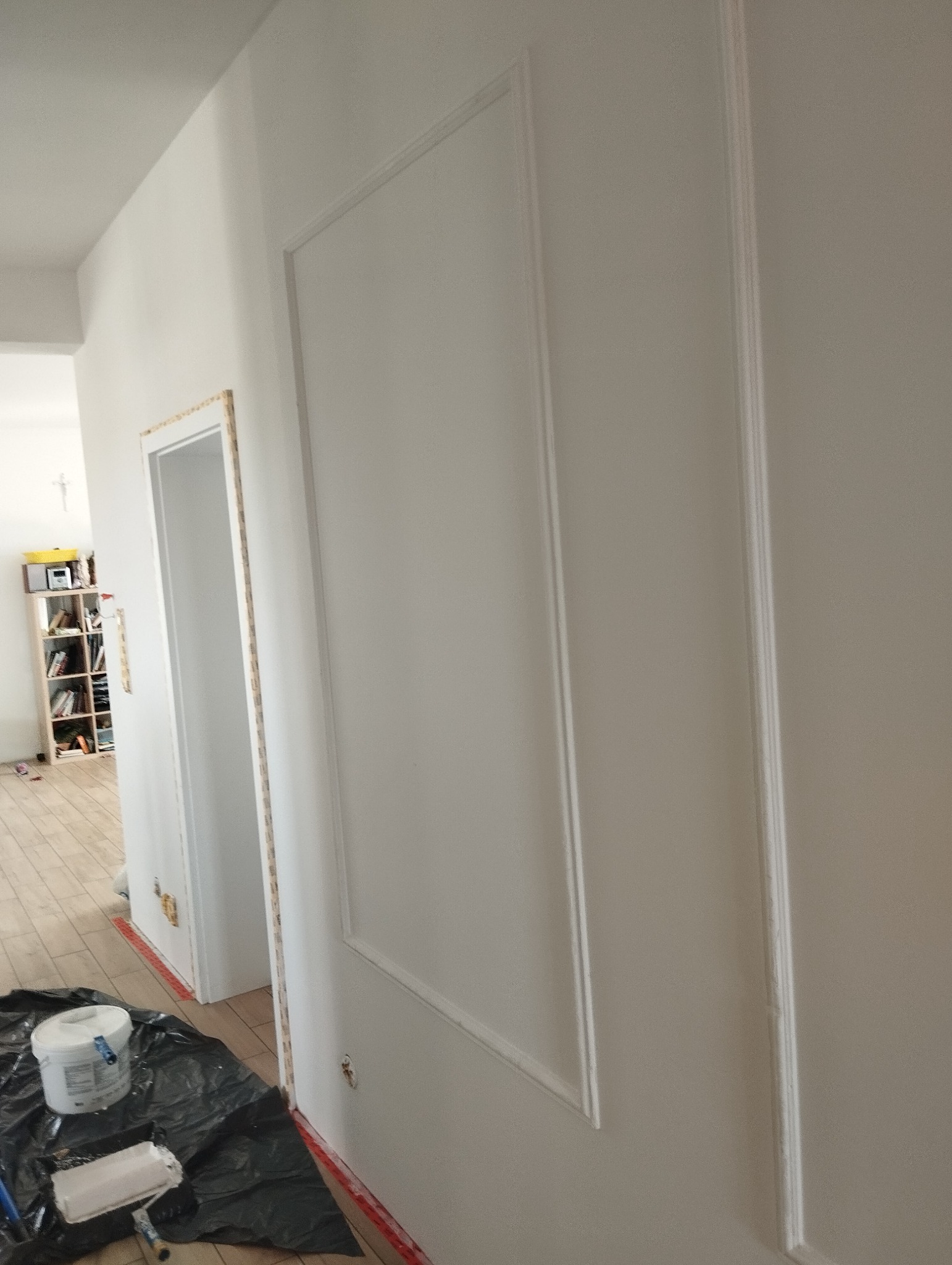 Białe ściany z dekoracyjnymi listwami po malowaniu, widoczny otwarty pokój i akcesoria malarskie. Minimalistyczne wykończenie wnętrza w jasnej tonacji.