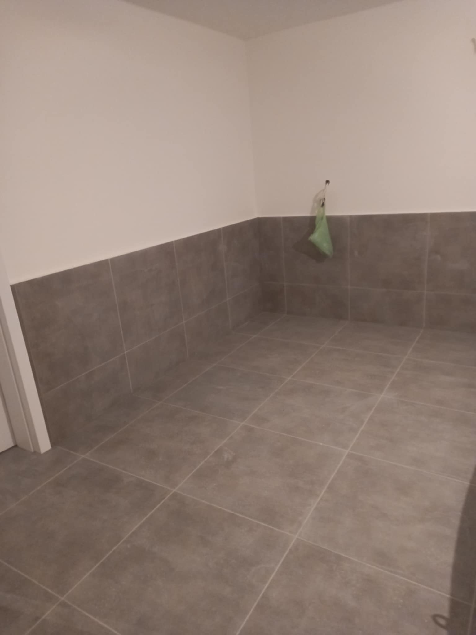 Wykończona łazienka z szarymi płytkami na podłodze i ścianach do połowy wysokości. Na ścianie wisi zielona torba foliowa. Widok z góry, neutralne oświetlenie.