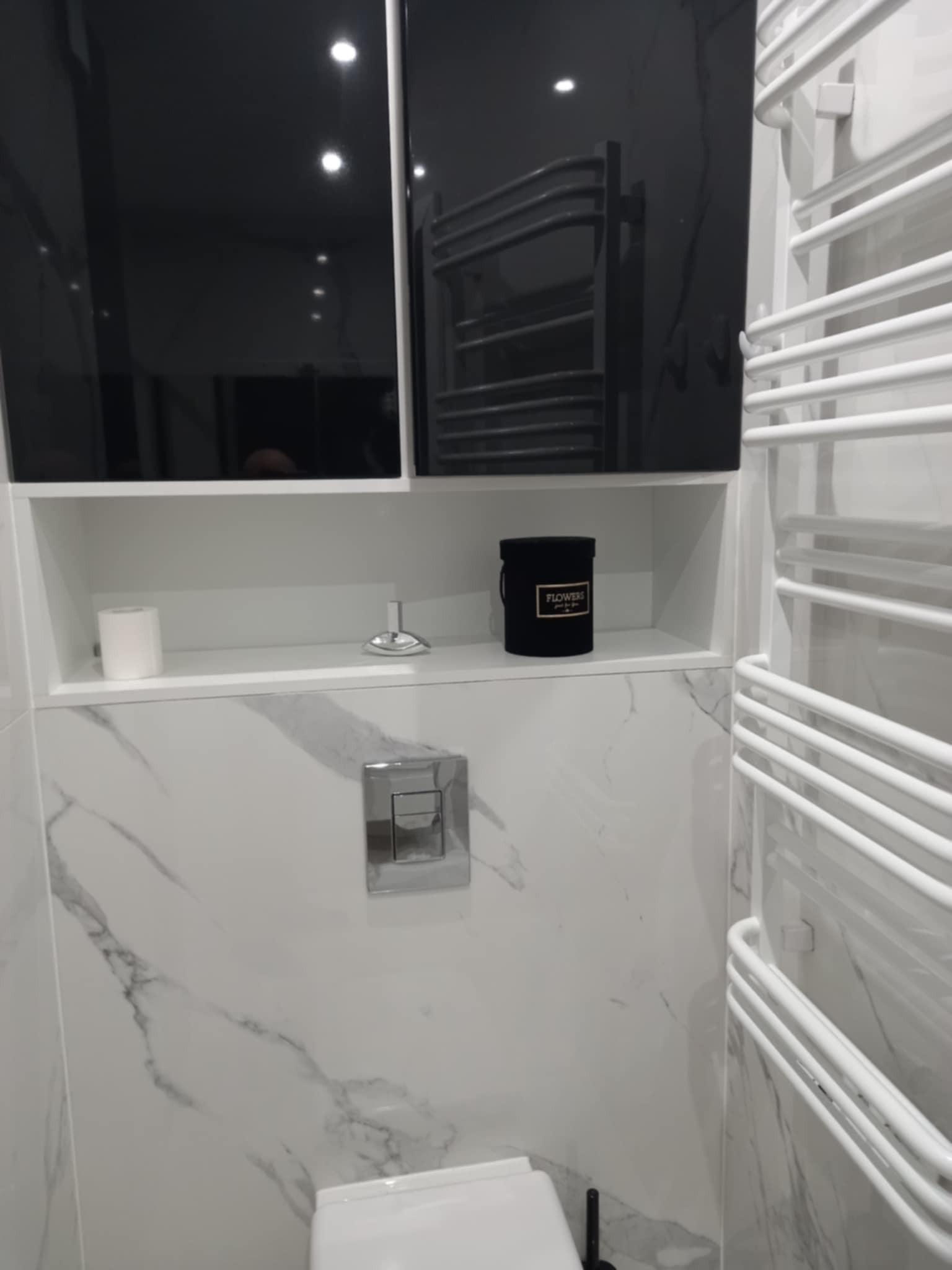 Nowoczesna łazienka z marmurowymi płytkami, czarnymi szafkami i białym grzejnikiem drabinkowym. Wnęka z dekoracjami: świeca, ozdoba i pudełko z kwiatami.