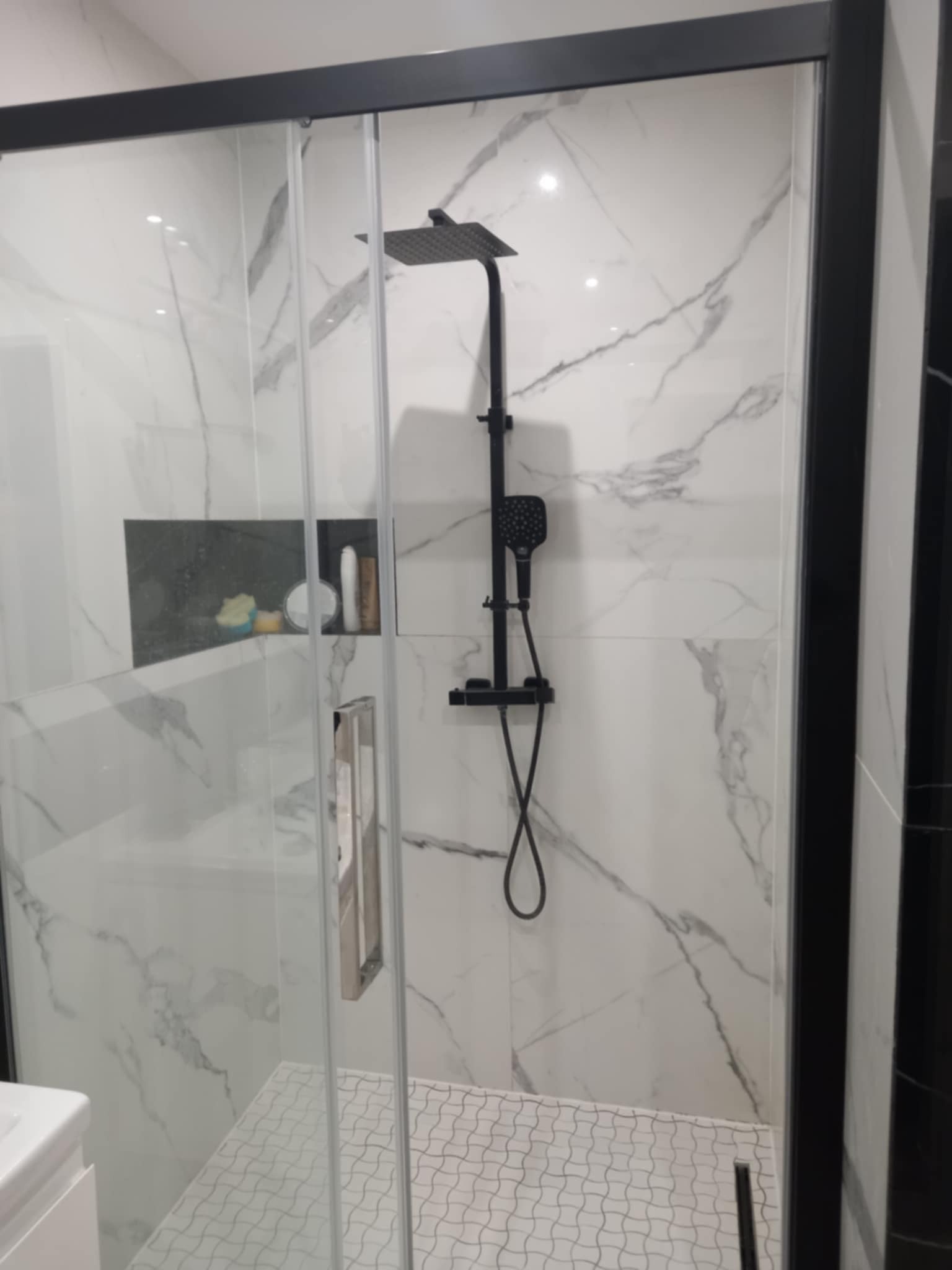 Nowoczesna kabina prysznicowa z czarną armaturą i białymi płytkami imitującymi marmur. Wnęka z kosmetykami. Biała mozaika podłogowa. Minimalistyczny design.