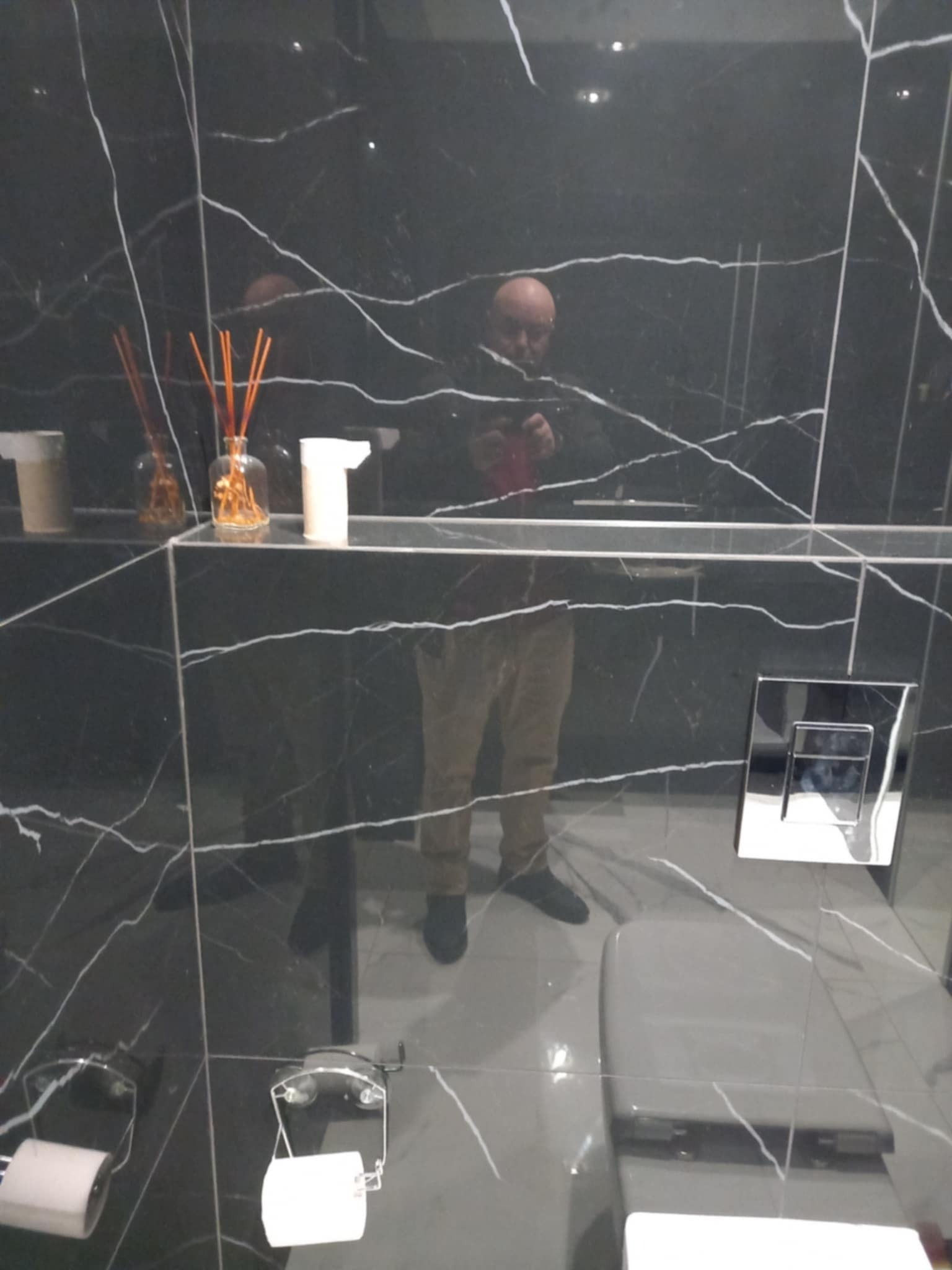 Elegancka łazienka wykończona czarnym marmurem z białymi żyłami. Widoczna toaleta, akcesoria łazienkowe i odbicie osoby w lustrze. Nowoczesny design.