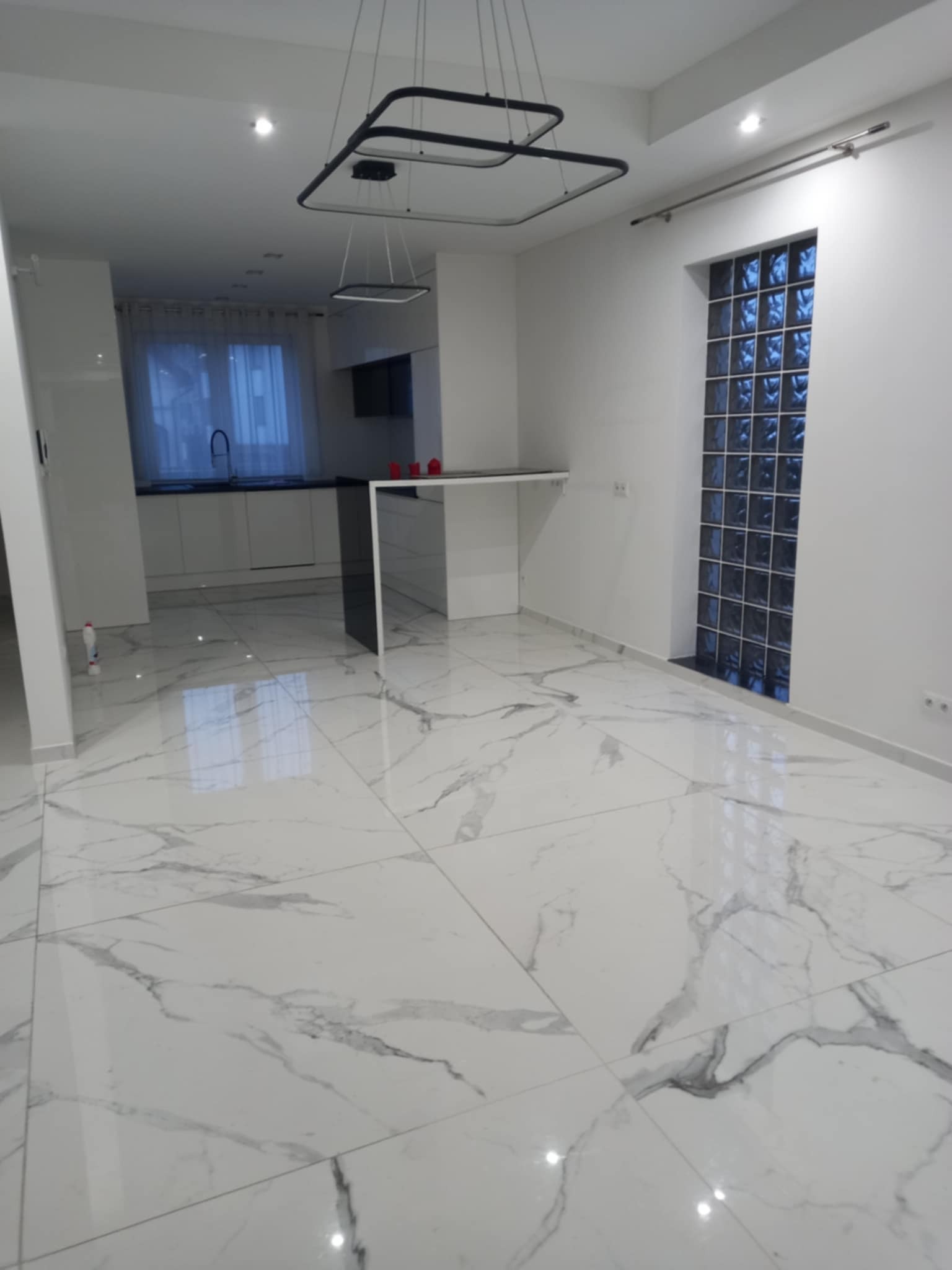 Wykończone wnętrze: biała marmurowa podłoga, nowoczesna kuchnia z czarnymi akcentami i szklana ściana z luksferów. Minimalistyczny design, jasne oświetlenie.