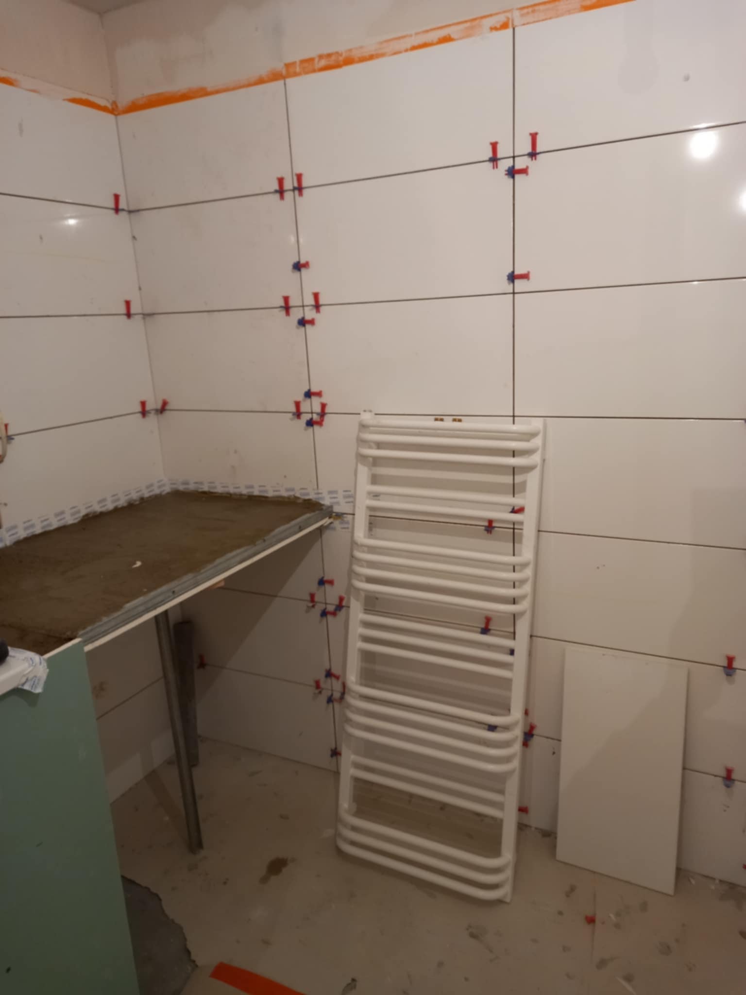 Białe płytki ścienne w trakcie układania, z krzyżykami dystansowymi. Widoczny grzejnik drabinkowy i przygotowane podłoże pod umywalkę. Prace wykończeniowe w łazience.