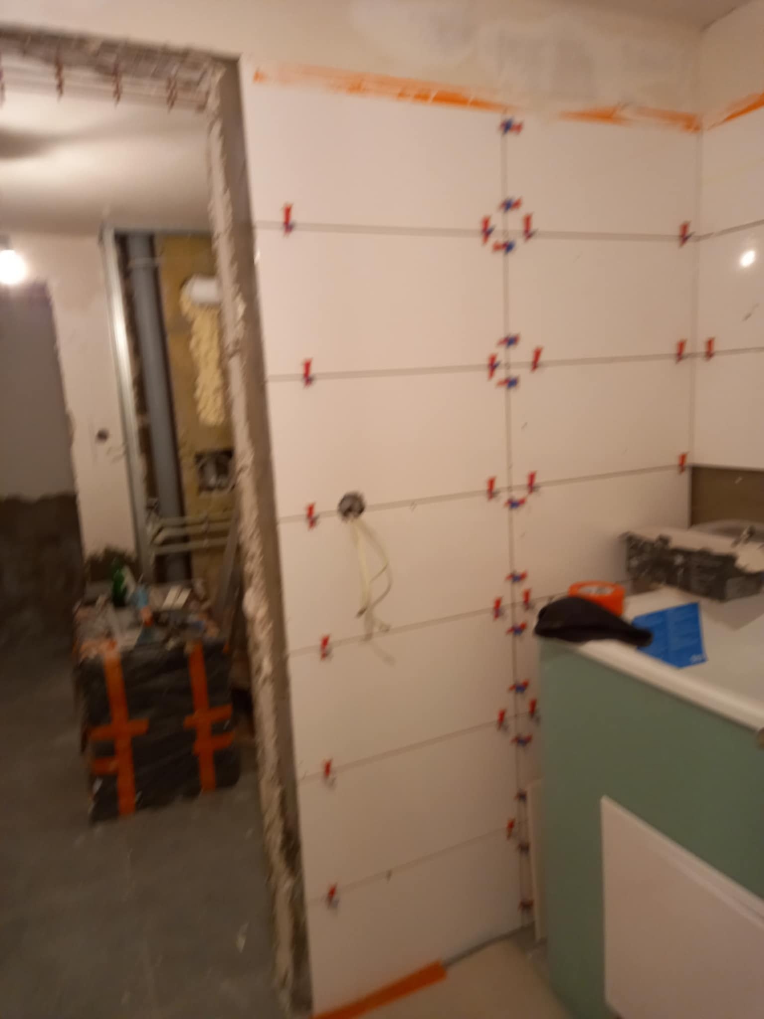 Białe płytki ścienne z krzyżykami dystansowymi w trakcie montażu. Widoczne narzędzia i materiały budowlane. W tle instalacja. Ujęcie z bliska, wnętrze w trakcie prac.