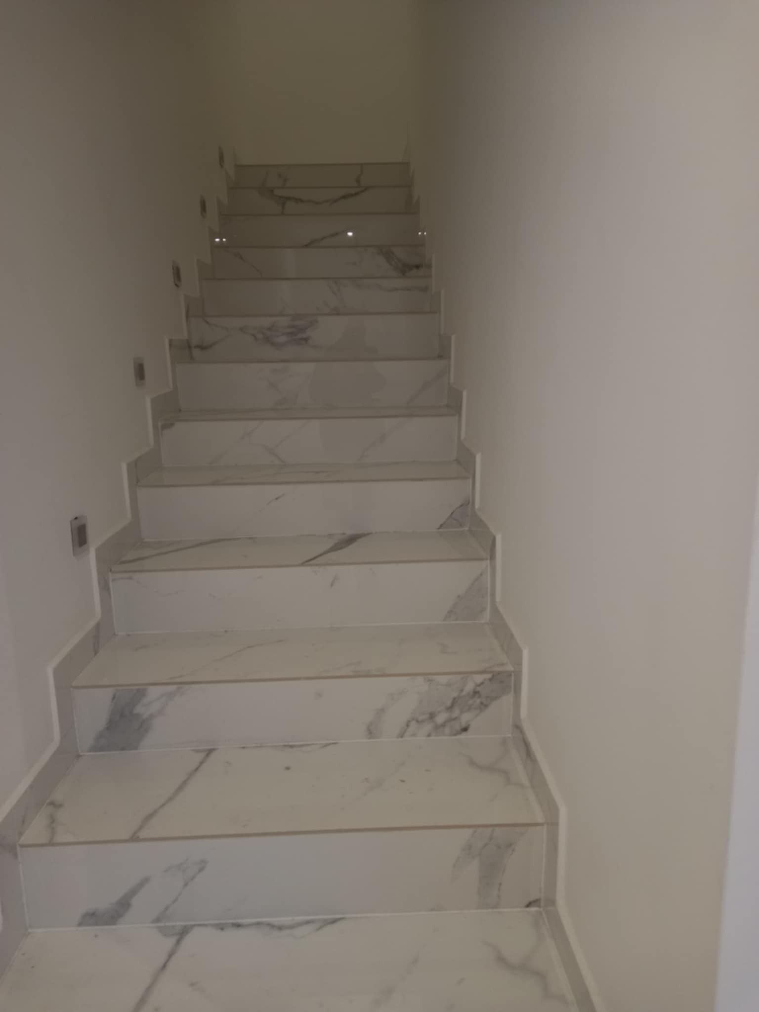 Schody wykończone białym marmurem z szarymi żyłami, oświetlone punktowymi lampami ściennymi. Widok z dołu, podkreślający elegancki design i precyzję wykonania.