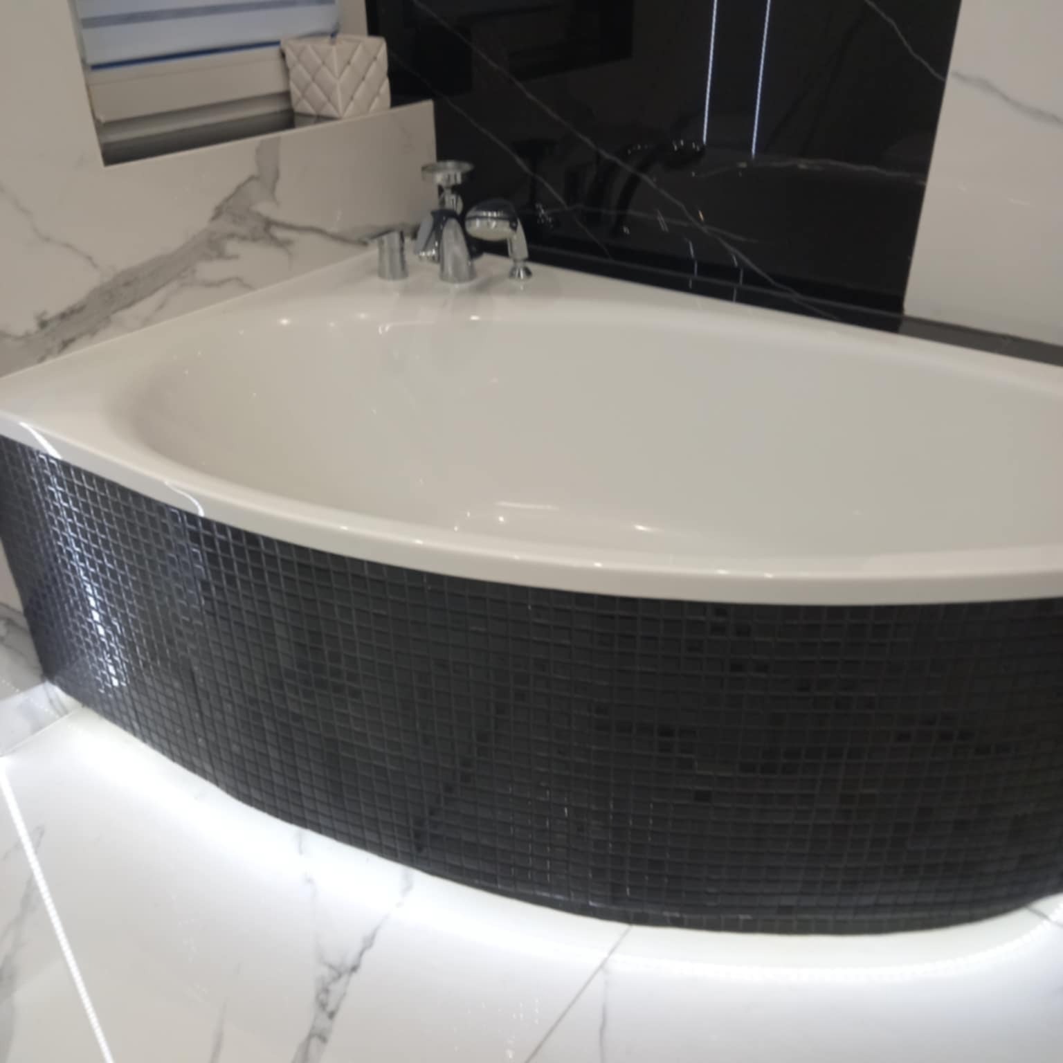 Nowoczesna, narożna wanna z czarną mozaiką i subtelnym podświetleniem LED, otoczona białym marmurem. Bateria chromowana. Minimalistyczny design łazienki.