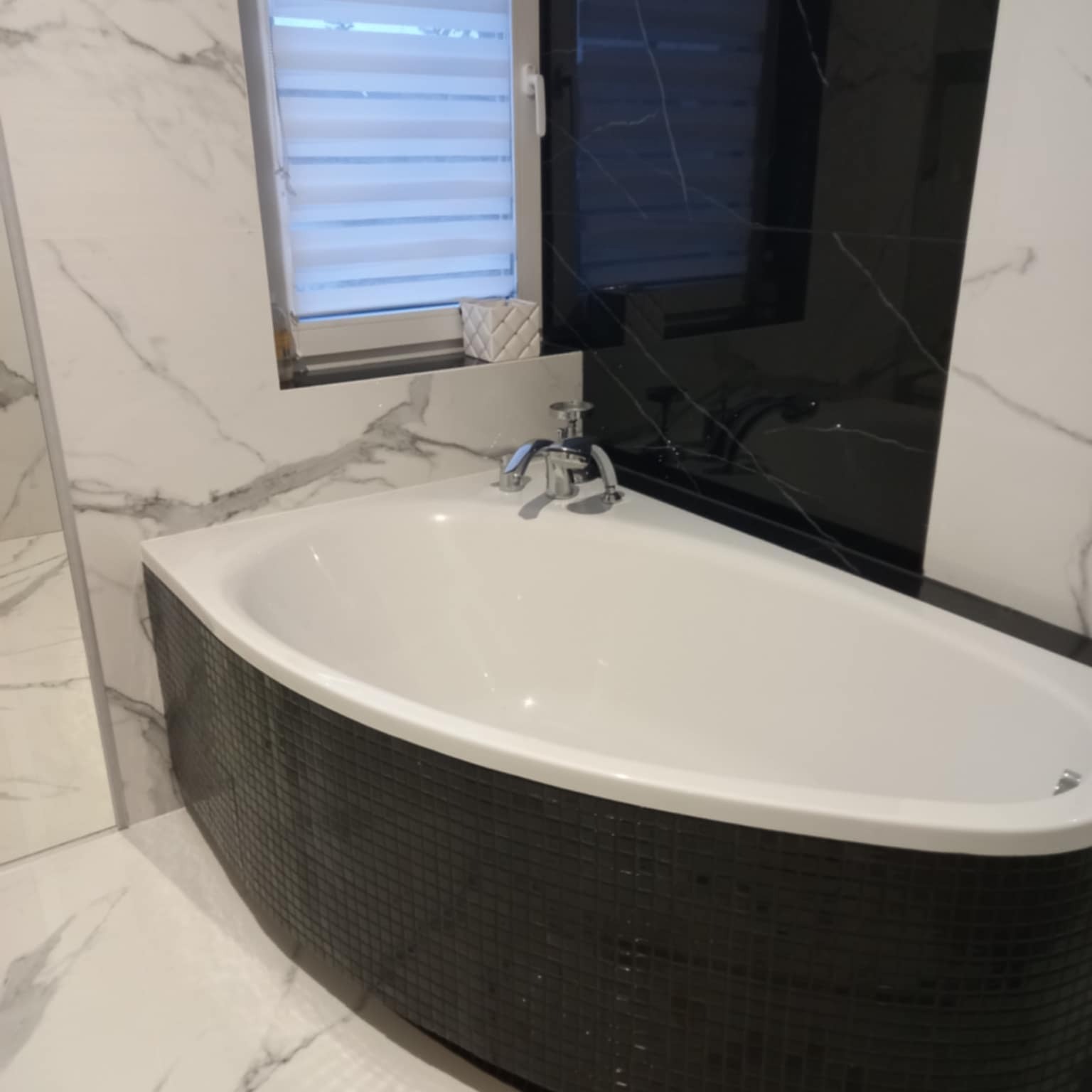 Nowoczesna łazienka z wolnostojącą wanną obłożoną czarną mozaiką, białe marmurowe płytki na ścianach i podłodze, okno z żaluzjami w tle.