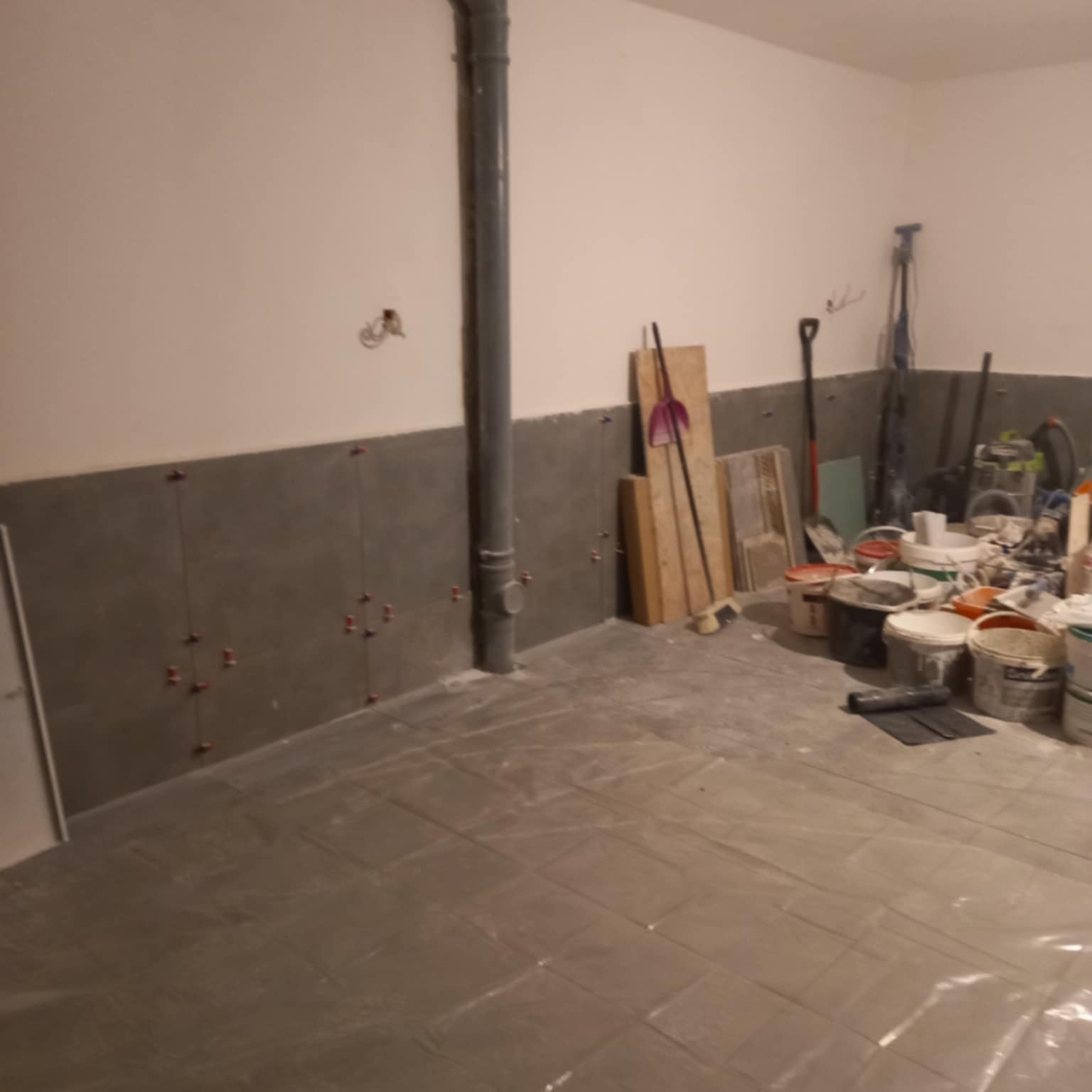 Wnętrze w trakcie układania płytek ściennych w szarym odcieniu. Na podłodze folia ochronna, widoczne narzędzia i materiały budowlane. Pionowa rura kanalizacyjna przy ścianie.