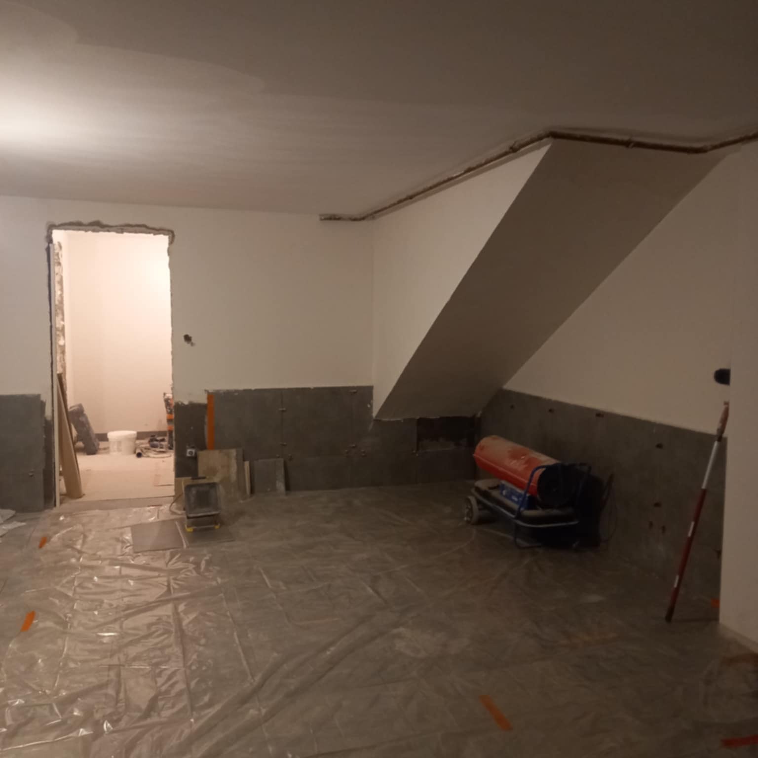 Wnętrze w trakcie prac wykończeniowych: ściany przygotowane pod gładzie, podłoga zabezpieczona folią, widoczny grzejnik budowlany i narzędzia.