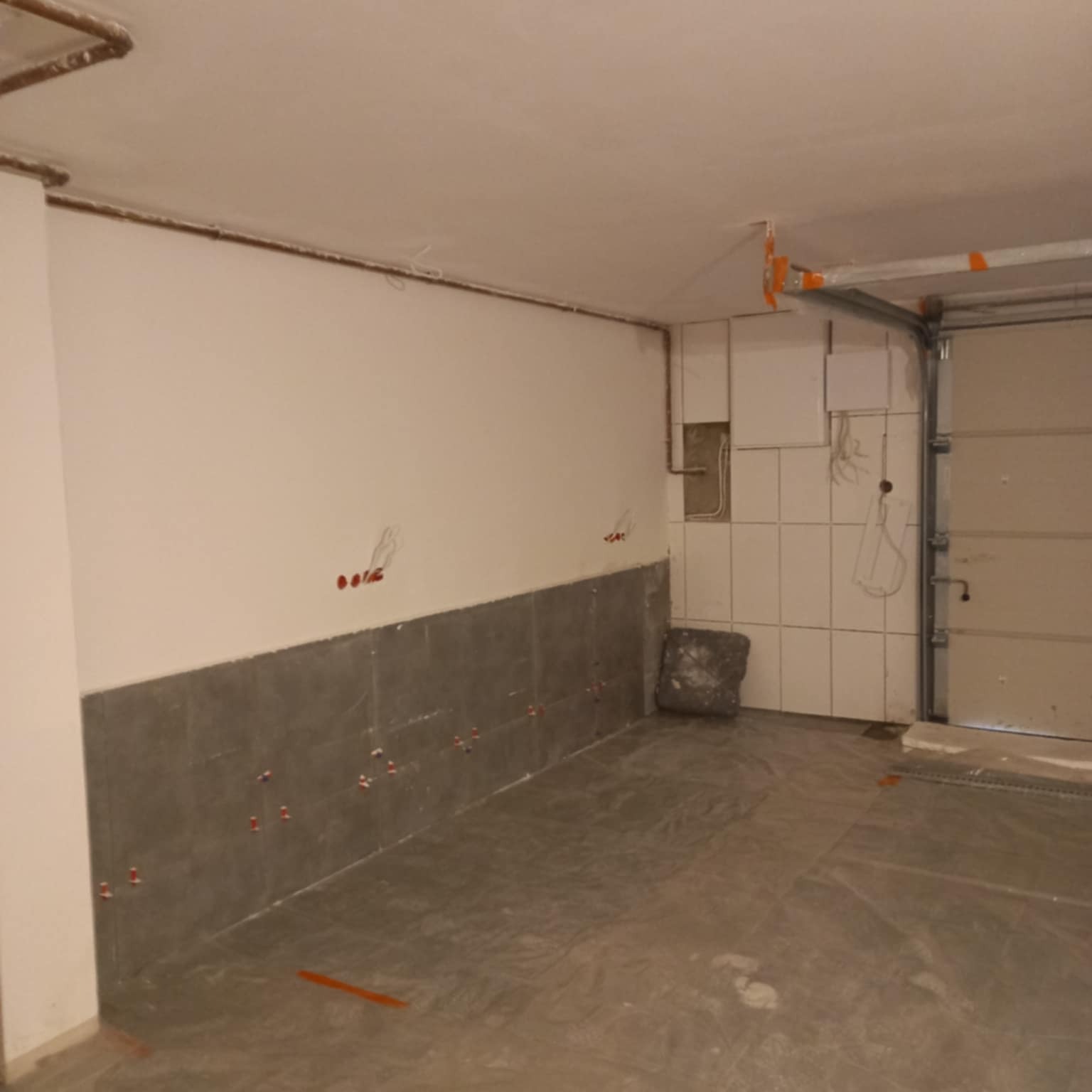 Wnętrze w trakcie układania płytek: szare płytki na podłodze i dolnej części ściany, białe płytki na górze ściany, widoczne krzyżyki dystansowe, garaż w tle.
