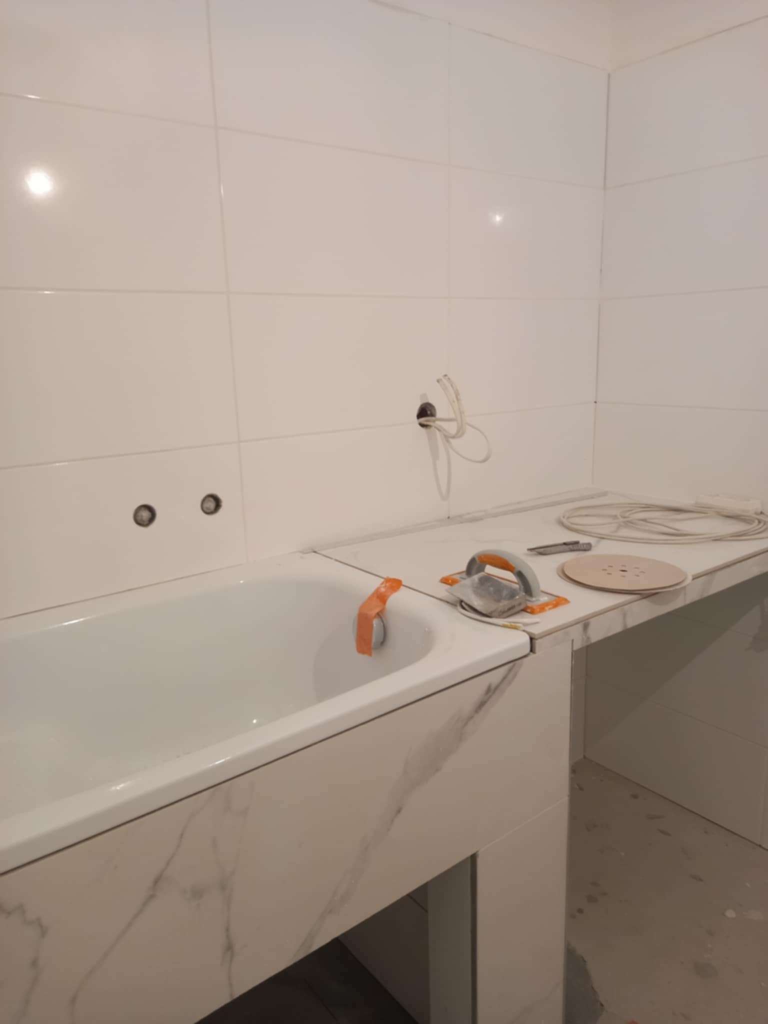 Biała wanna z marmurowym wykończeniem, obok narzędzia glazurnicze i kable. Ściany wyłożone białymi płytkami. Widoczne gniazdka i otwór na kable.