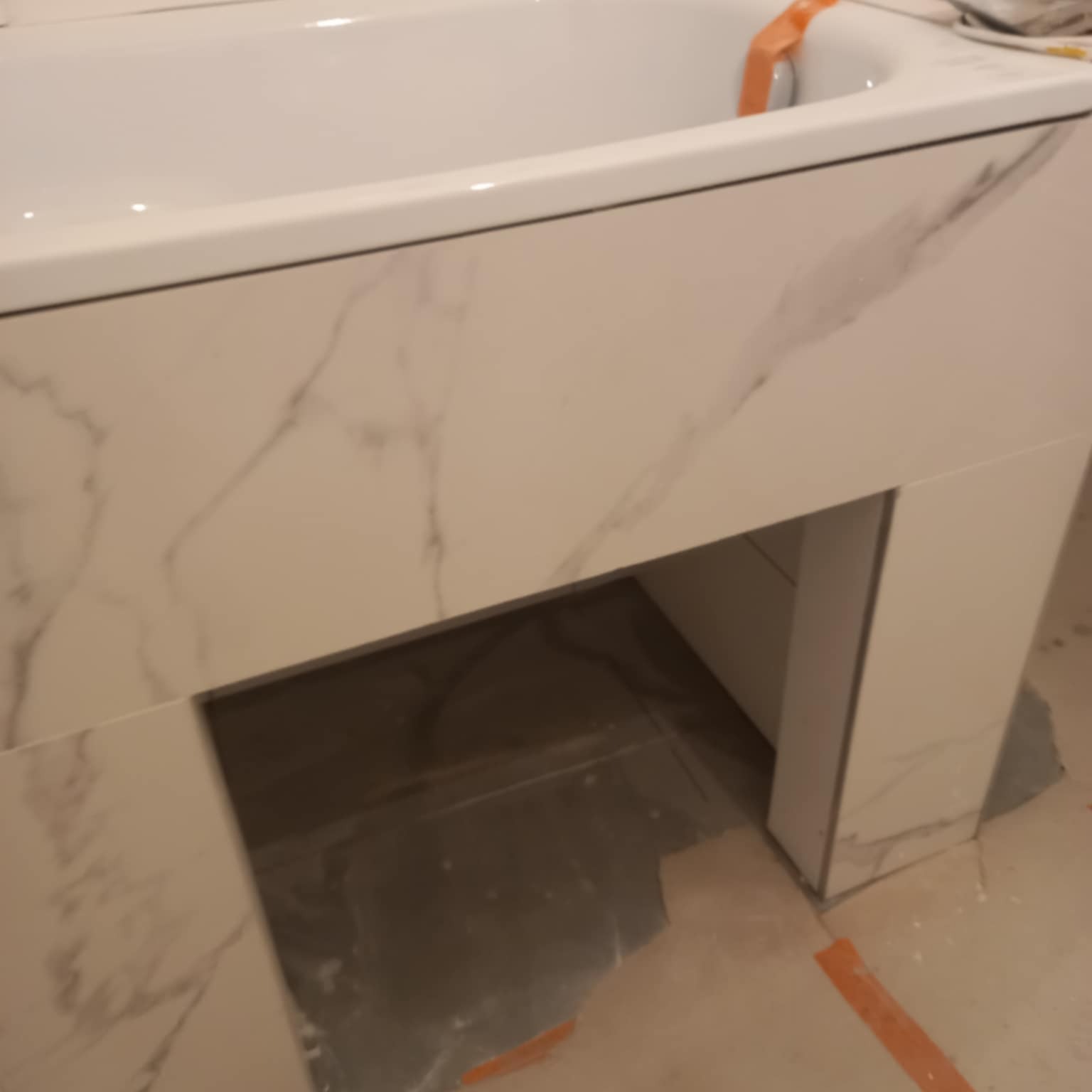 Obudowa wanny wykończona płytkami imitującymi marmur, z widocznym otworem rewizyjnym. Detal wykończenia łazienki w trakcie remontu, widoczne zabezpieczenia.