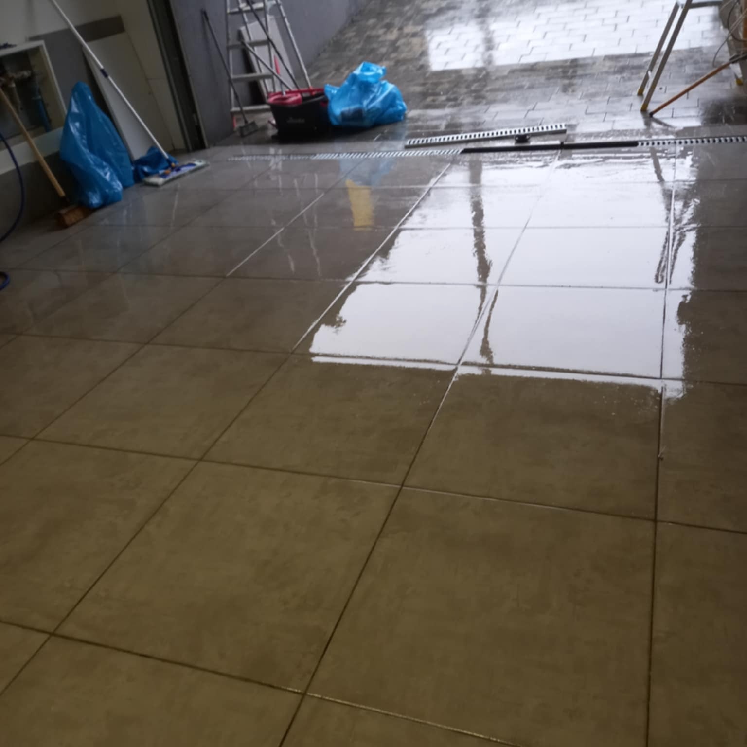 Błyszcząca, mokra podłoga z płytek po umyciu, z widocznymi narzędziami do sprzątania i drabiną w tle. Świeżo umyta posadzka z refleksami światła.