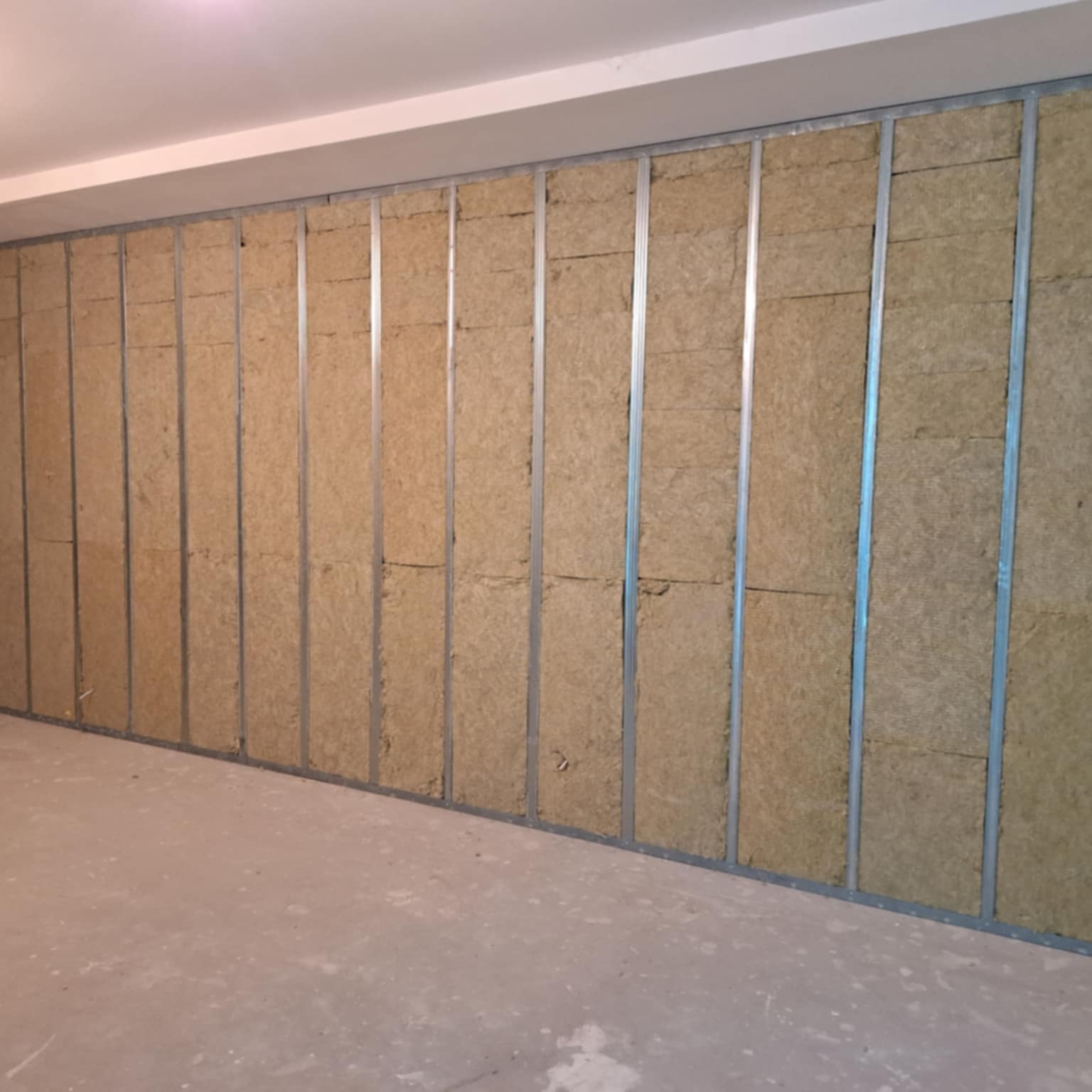 Ściana z metalowym stelażem wypełnionym wełną mineralną, przygotowana do montażu płyt karton-gips. Widoczne profile i izolacja termiczna w surowym wnętrzu.