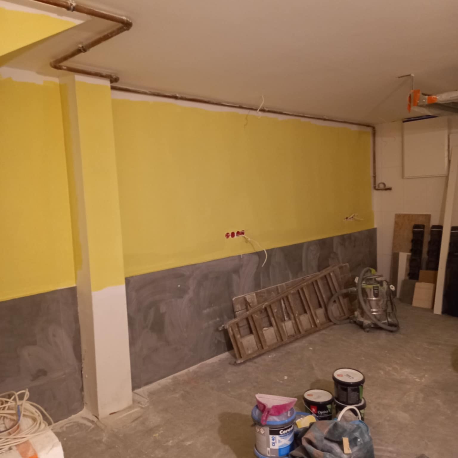 Żółta ściana w trakcie malowania, widoczne puszki z farbą, drabina i odkurzacz. Dolna część ściany pomalowana na szaro. Instalacja elektryczna na ścianie.