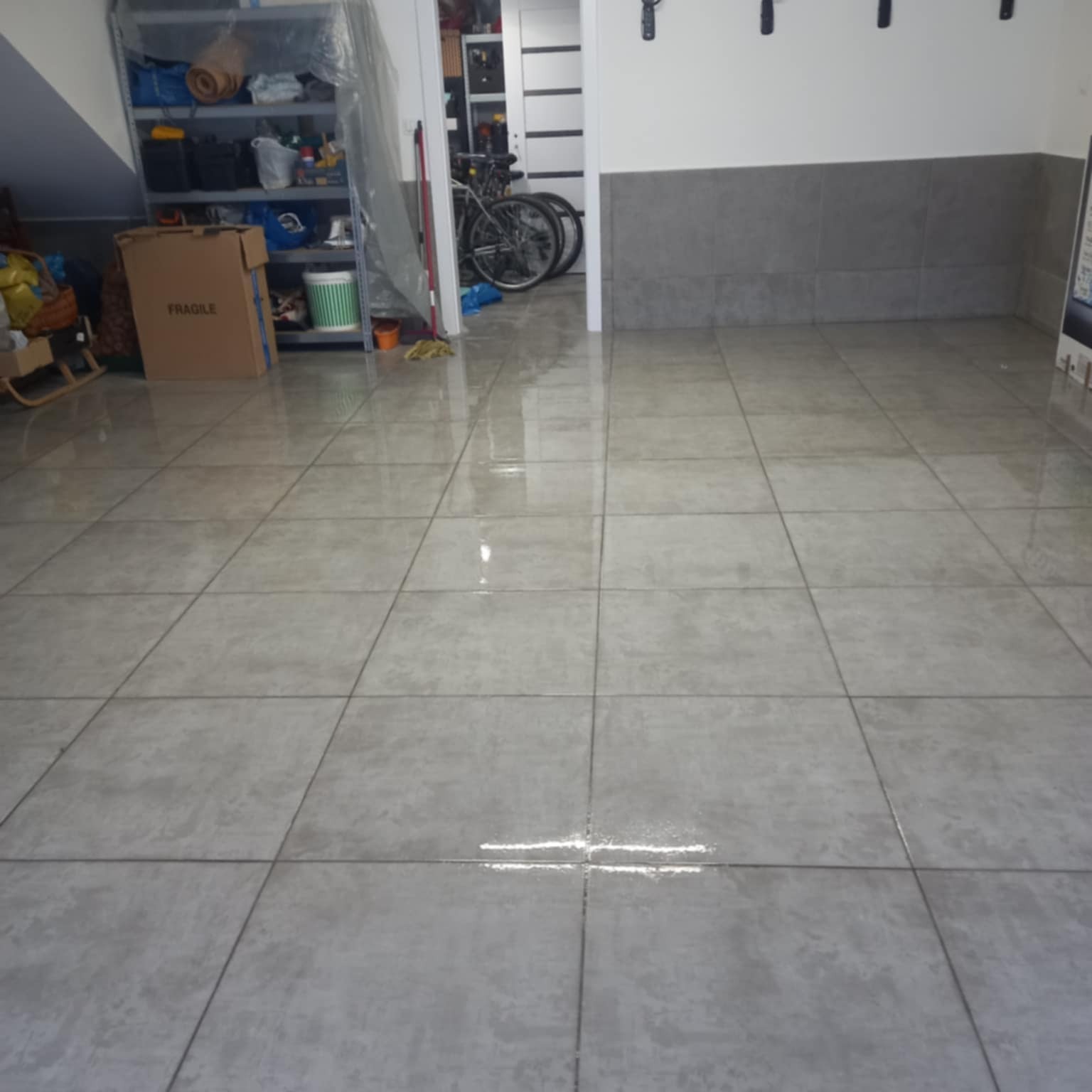 Wykończona podłoga z szarych, kwadratowych płytek w pomieszczeniu z szarą lamperią. Widoczne regały i rowery w tle. Świeżo położone płytki, odbijające światło.