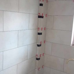 Daniel Sobota - Ściana w trakcie układania płytek w łazience, widoczne krzyżyki dystansowe i czarne elementy stabilizujące narożniki. Jasne, prostokątne płytki ułożone poziomo.