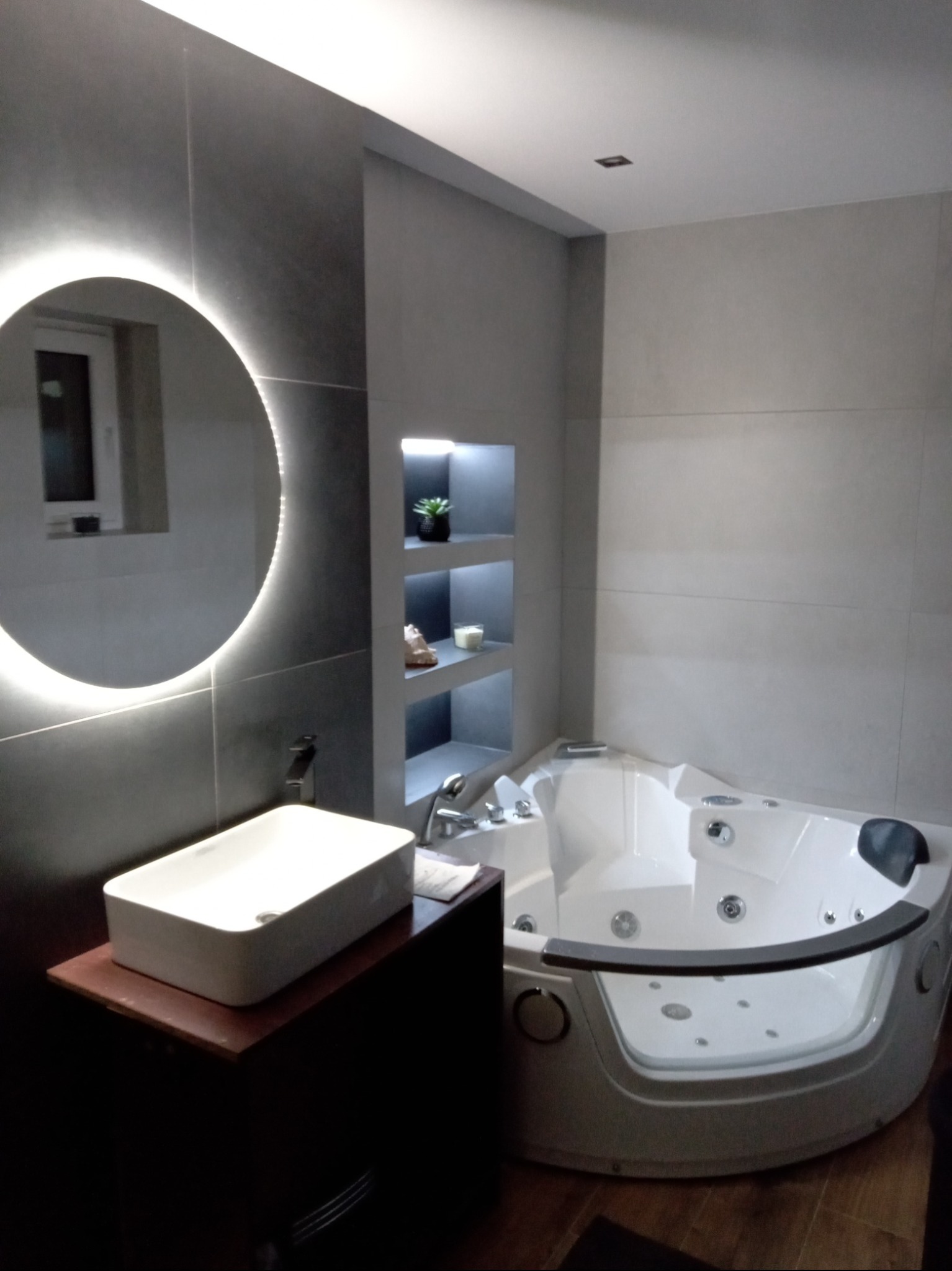 Nowoczesna łazienka z okrągłym lustrem LED, białą umywalką na ciemnej szafce i narożną wanną z hydromasażem. Szare płytki na ścianach i wnęka z półkami.