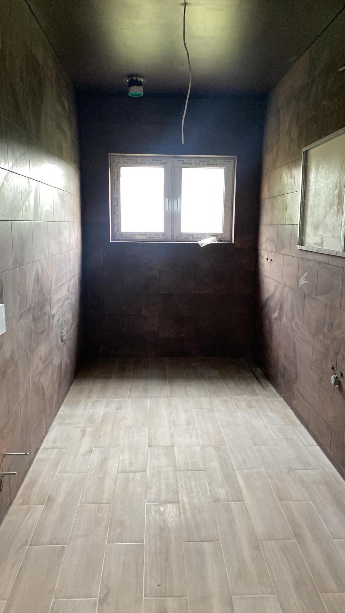 Wykończona łazienka z brązowymi płytkami na ścianach i beżowymi na podłodze. Widoczne okno i wystający kabel. Minimalistyczny design, surowe wykończenie.