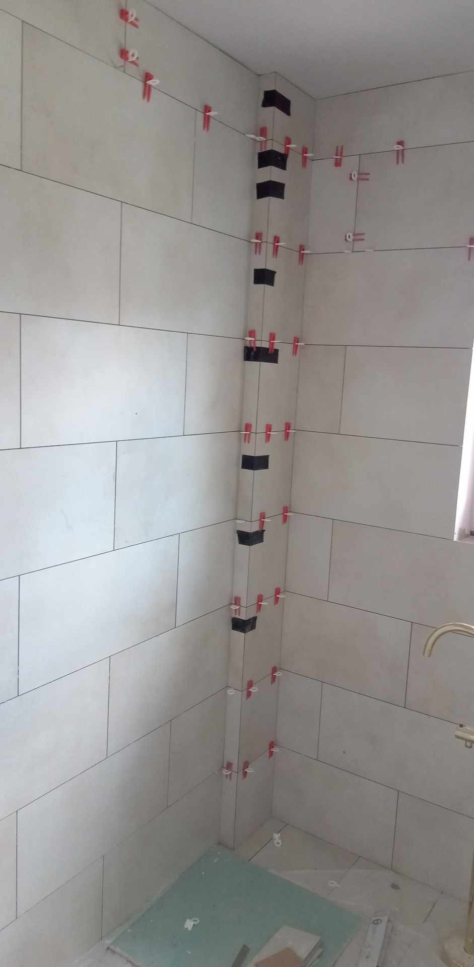 Ściana w trakcie układania płytek w łazience, widoczne krzyżyki dystansowe i czarne elementy stabilizujące narożniki. Jasne, prostokątne płytki ułożone poziomo.