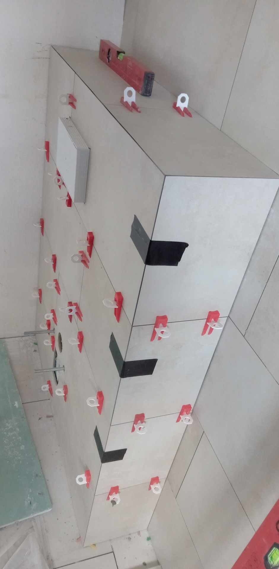 Świeżo ułożone, jasne płytki na ścianie, z użyciem systemu poziomowania (czerwone klipsy i białe kliny). Widoczny poziomica i fragment czarnej taśmy.