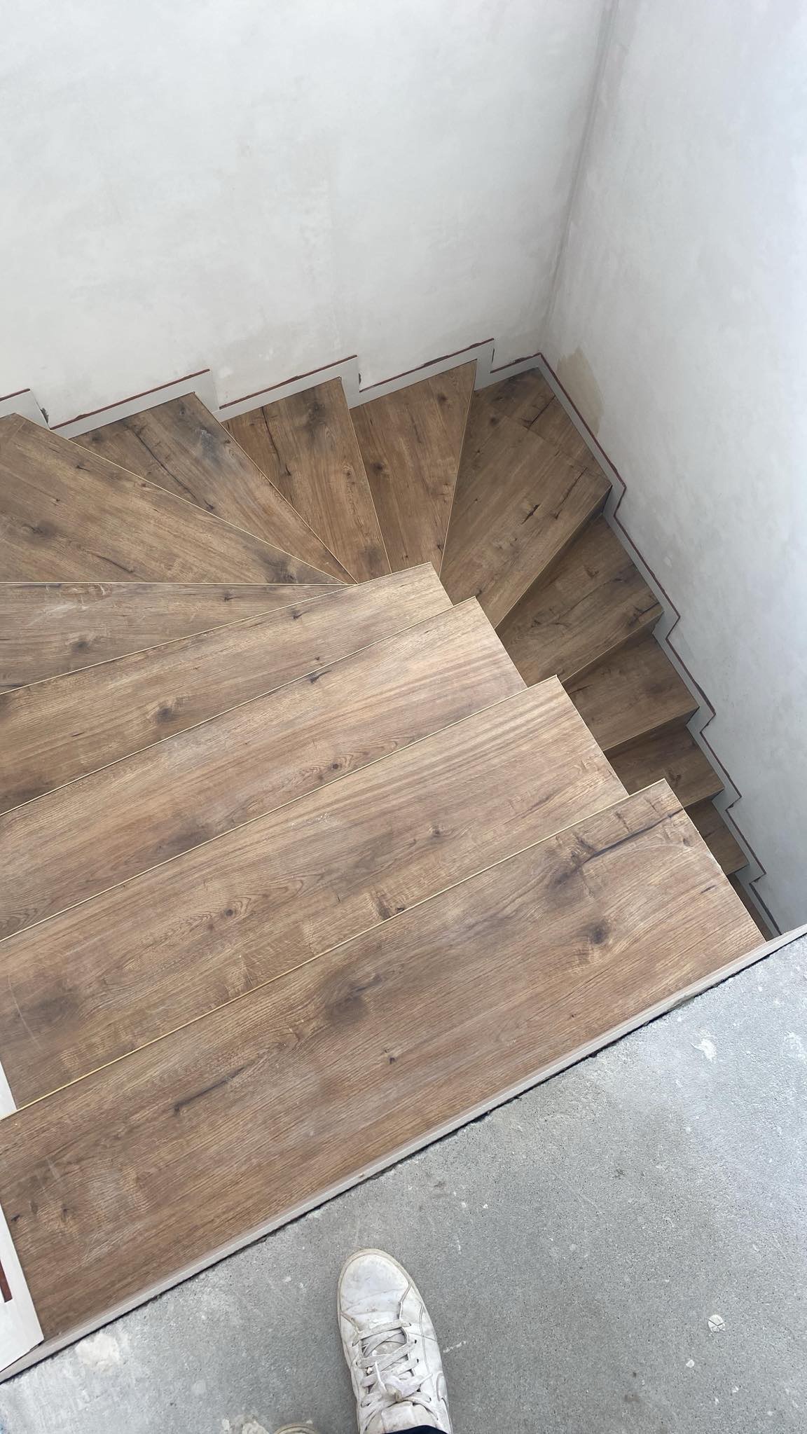 Spiralne schody wyłożone panelami imitującymi drewno, widok z góry, z fragmentem buta w dolnym rogu. Ściany w surowym stanie, przygotowane do wykończenia.