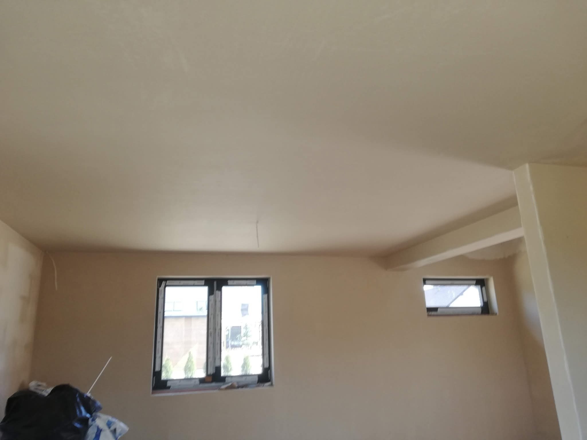 Wnętrze po szpachlowaniu: gładkie ściany w beżowym odcieniu, dwa okna z czarnymi ramami, widoczne worki z materiałami budowlanymi w rogu pomieszczenia.