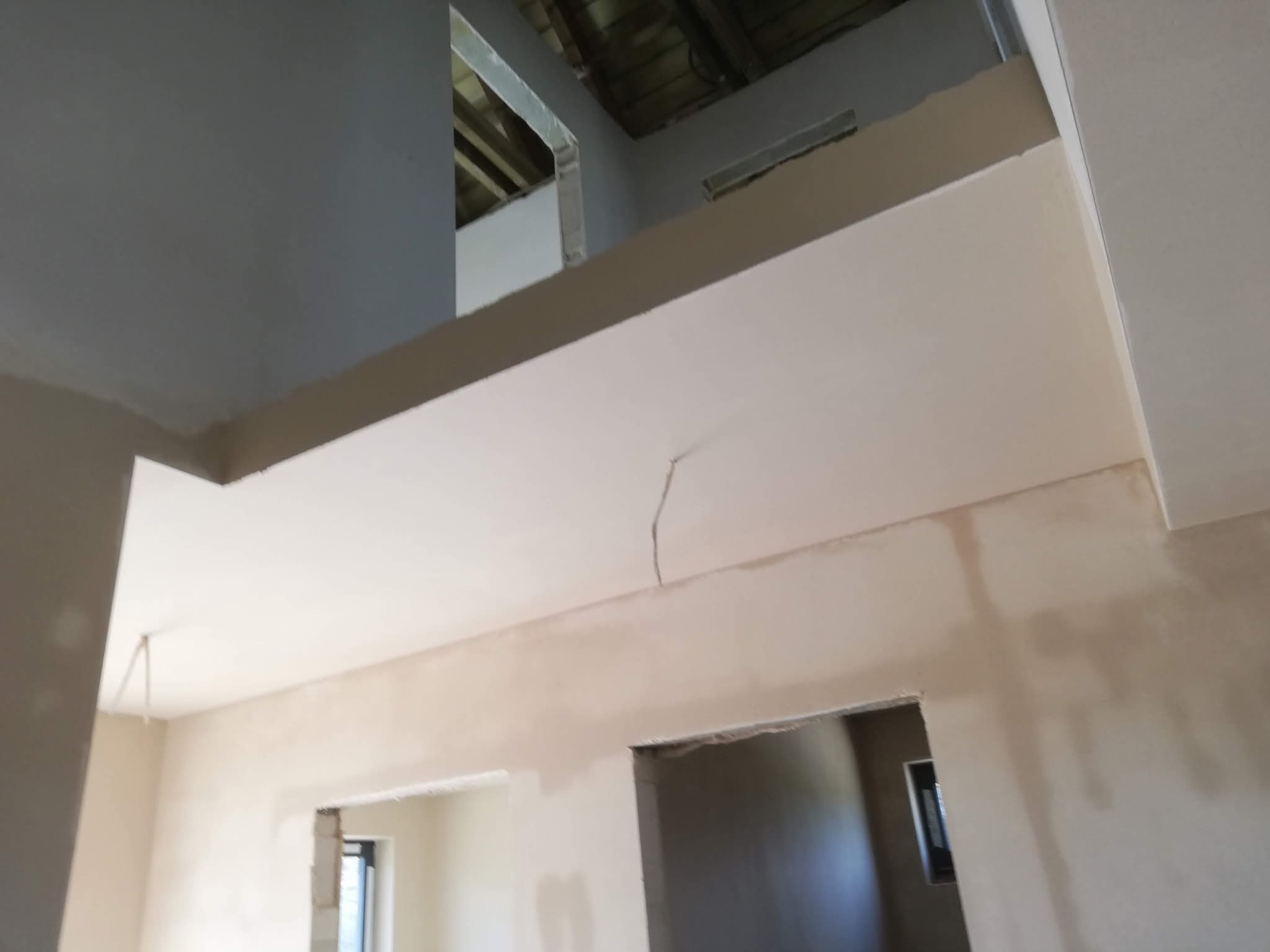 Surowe wnętrze w trakcie prac wykończeniowych. Widoczne ściany po szpachlowaniu, sufit z widoczną konstrukcją. Ujęcie z dołu, podkreślające przestrzeń i etap prac.