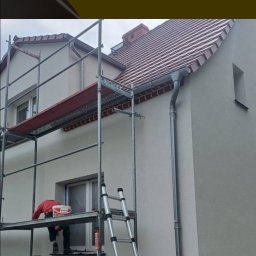 PM-Dekor Paweł Miklas - Ocieplanie elewacji domu jednorodzinnego. Pracownik na rusztowaniu, widoczne narzędzia i materiały budowlane. Świeżo otynkowana ściana, nowy system rynnowy.