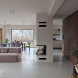 PM-Dekor Paweł Miklas - Nowoczesne wnętrze z szarym betonem architektonicznym na podłodze, minimalistycznym kominkiem i widokiem na ogród przez duże okno. W tle widoczna kuchnia.