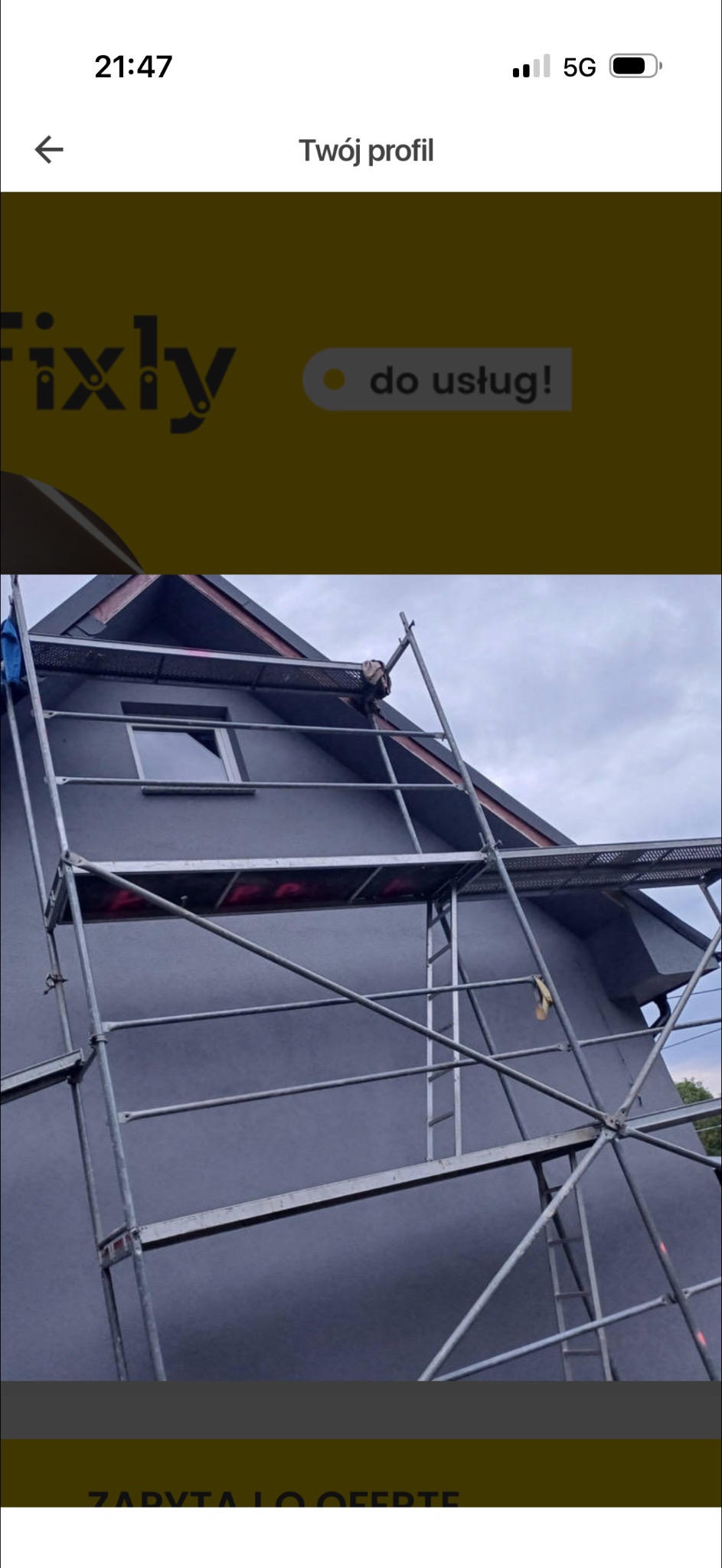 Szara fasada domu jednorodzinnego z metalowym rusztowaniem. Widoczne okno i fragment dachu. Pochmurne niebo w tle. Ujęcie z dołu.