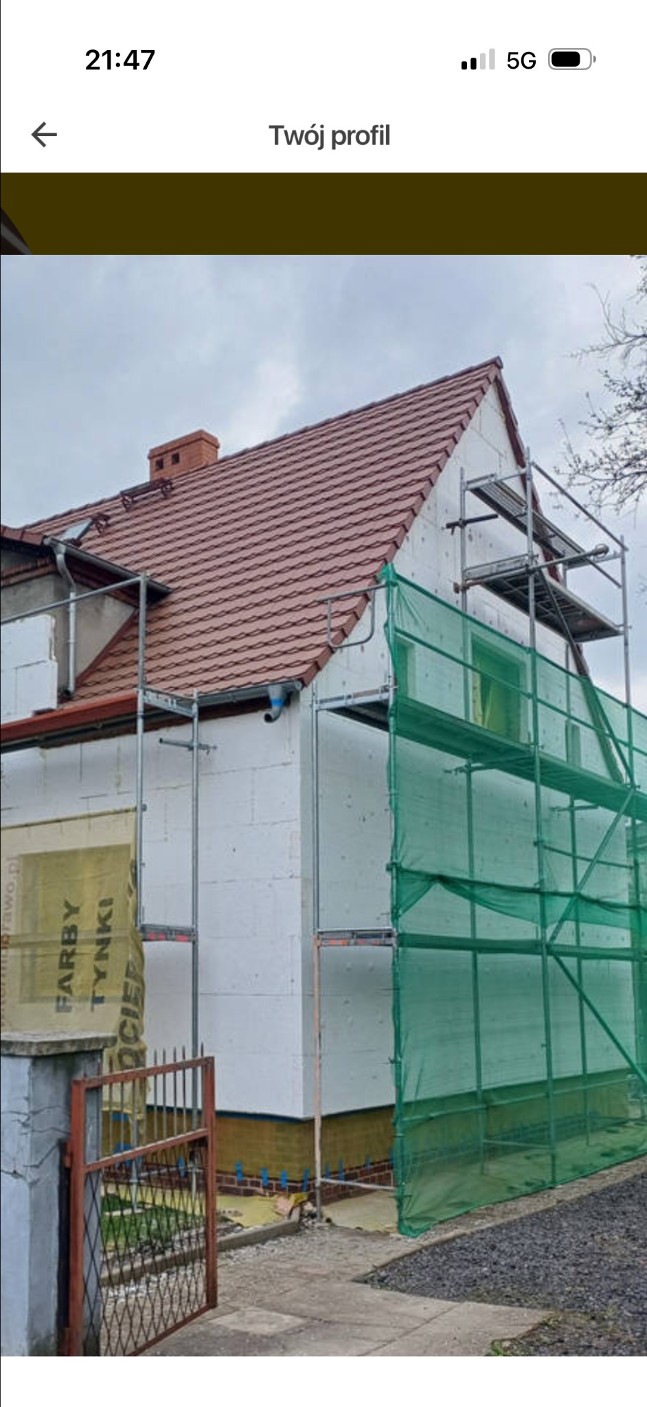 Ocieplanie domu styropianem, widoczne rusztowania i siatka ochronna. Częściowo ocieplona ściana z widoczną reklamą farb i tynków. Dach pokryty czerwoną dachówką.