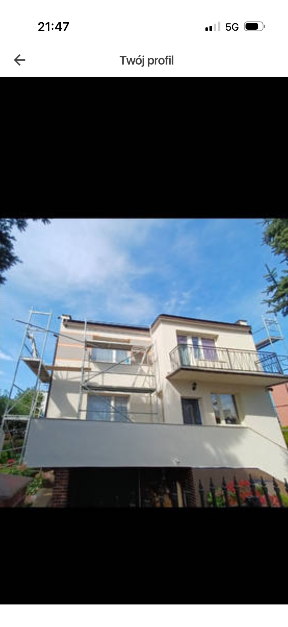 Dwupiętrowy dom z balkonem w trakcie prac wykończeniowych elewacji. Widoczne rusztowania i fragmenty ocieplenia. Błękitne niebo w tle.