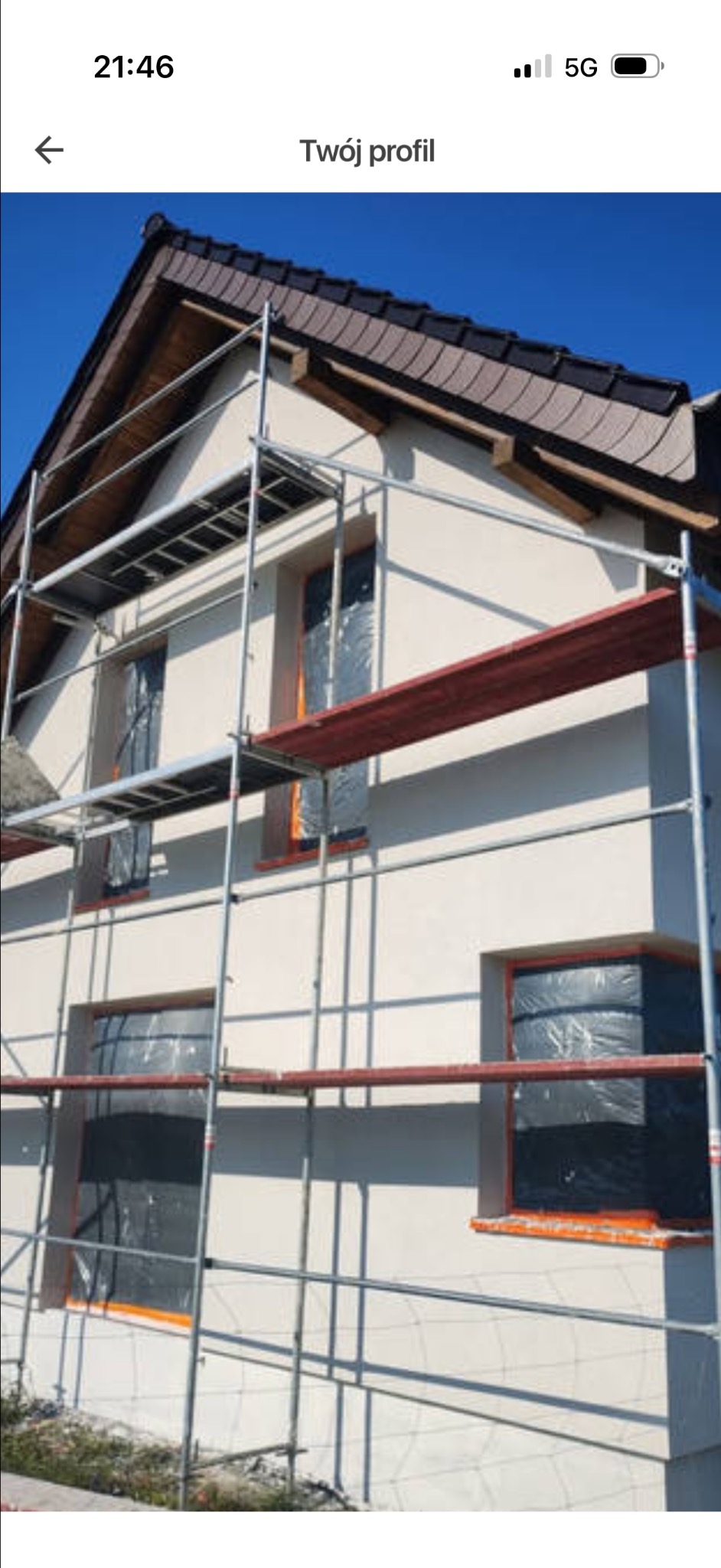 Ocieplanie elewacji domu jednorodzinnego. Okna zabezpieczone folią, rusztowanie wzdłuż ściany. Widok na fragment dachu z brązową dachówką.