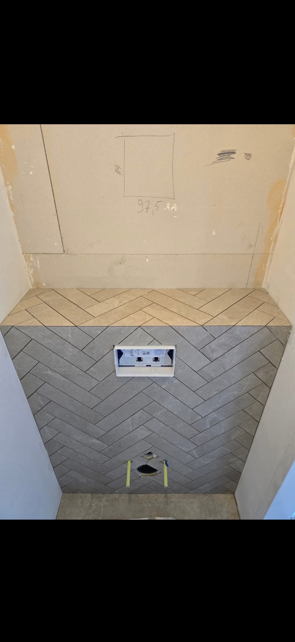 Ściana w łazience z szarą i beżową glazurą ułożoną w jodełkę, widoczne otwory na rury i instalację podtynkową. Na górze ściany widoczne notatki i szkic.