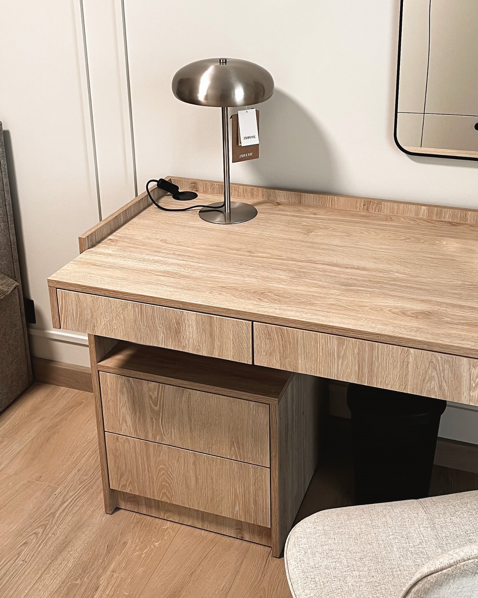 Drewniane biurko z szufladami i lampką w stylu retro, uzupełnione o lustro z czarną ramą. Jasne wnętrze, minimalistyczny design, widoczny fragment krzesła.