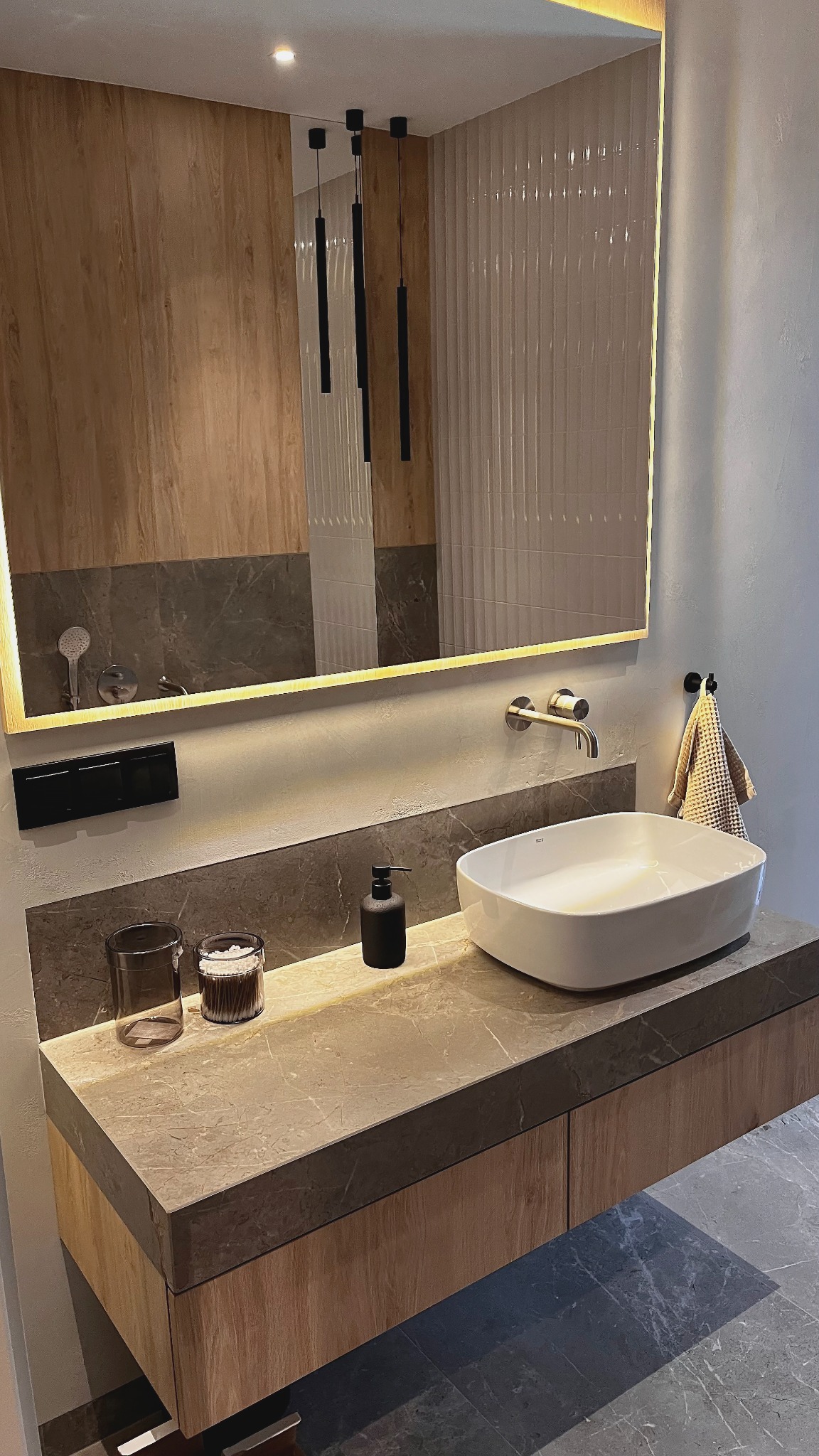 Nowoczesna łazienka z podświetlanym lustrem, marmurowym blatem i drewnianą szafką. Minimalistyczny design, eleganckie dodatki i stonowana kolorystyka.